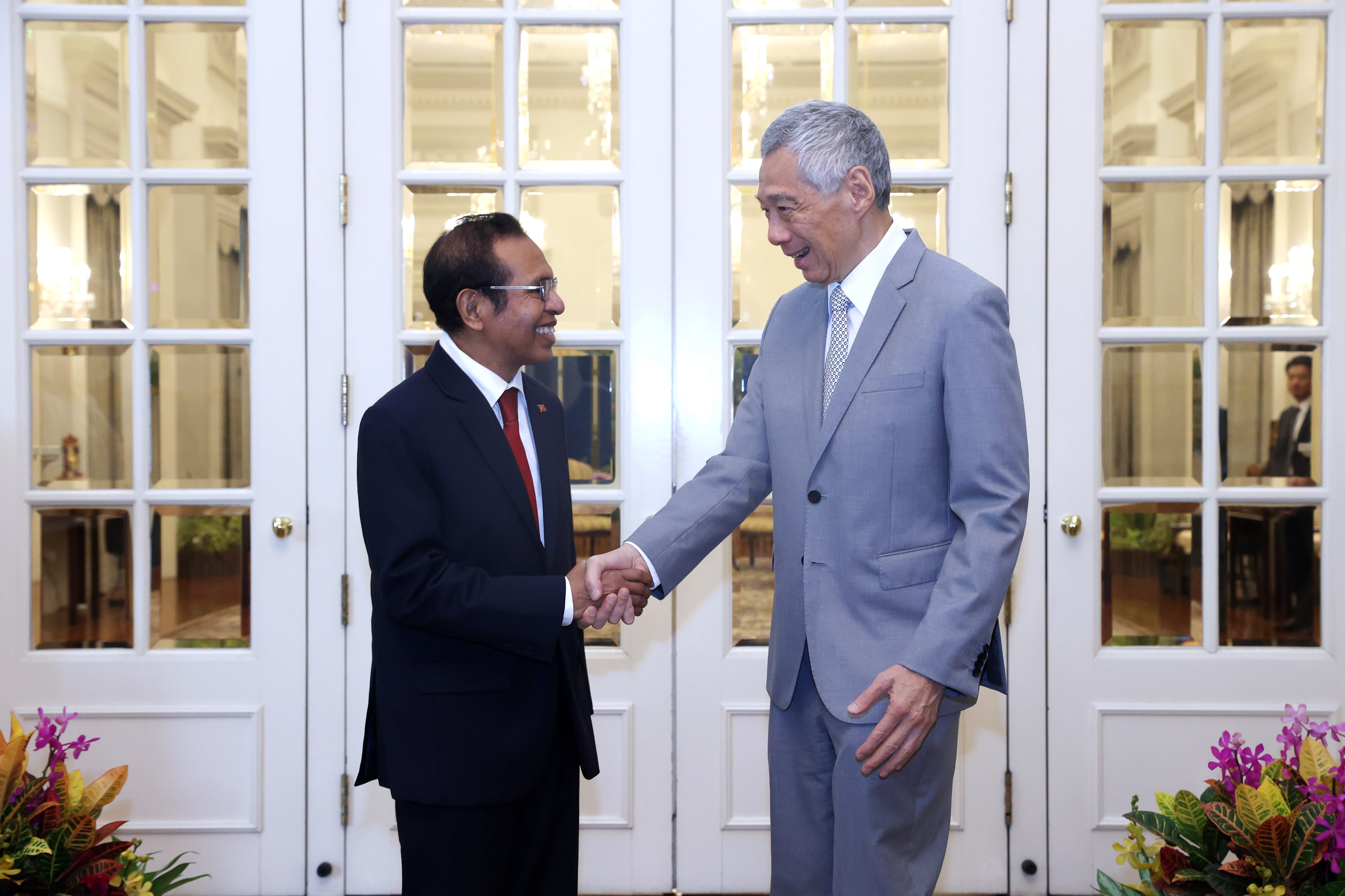 Two men in suits shake hands in front of glass-paneled doors.