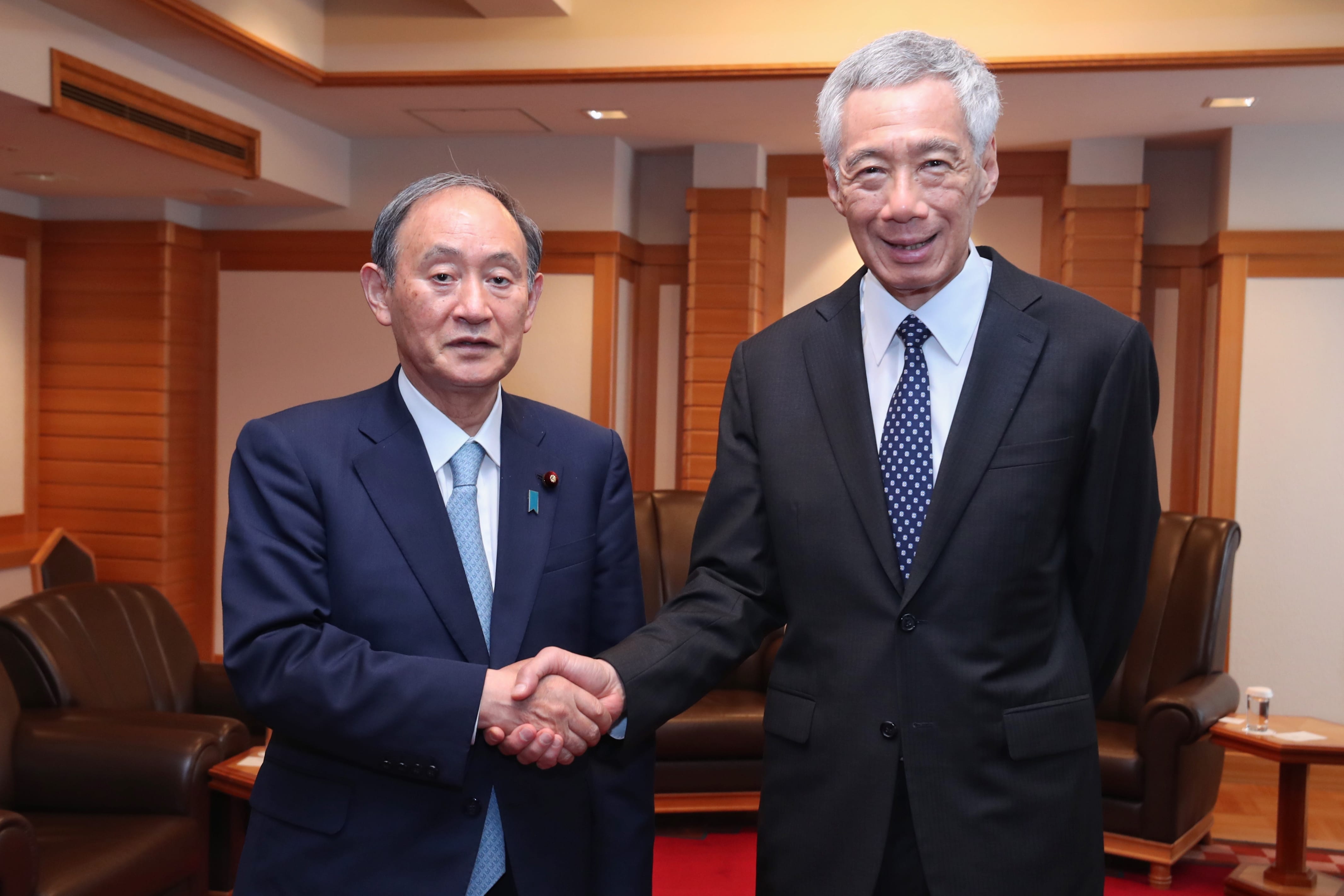 Lee Hsien Loong and Yoshihide Suga shake hands in a wood paneled room.