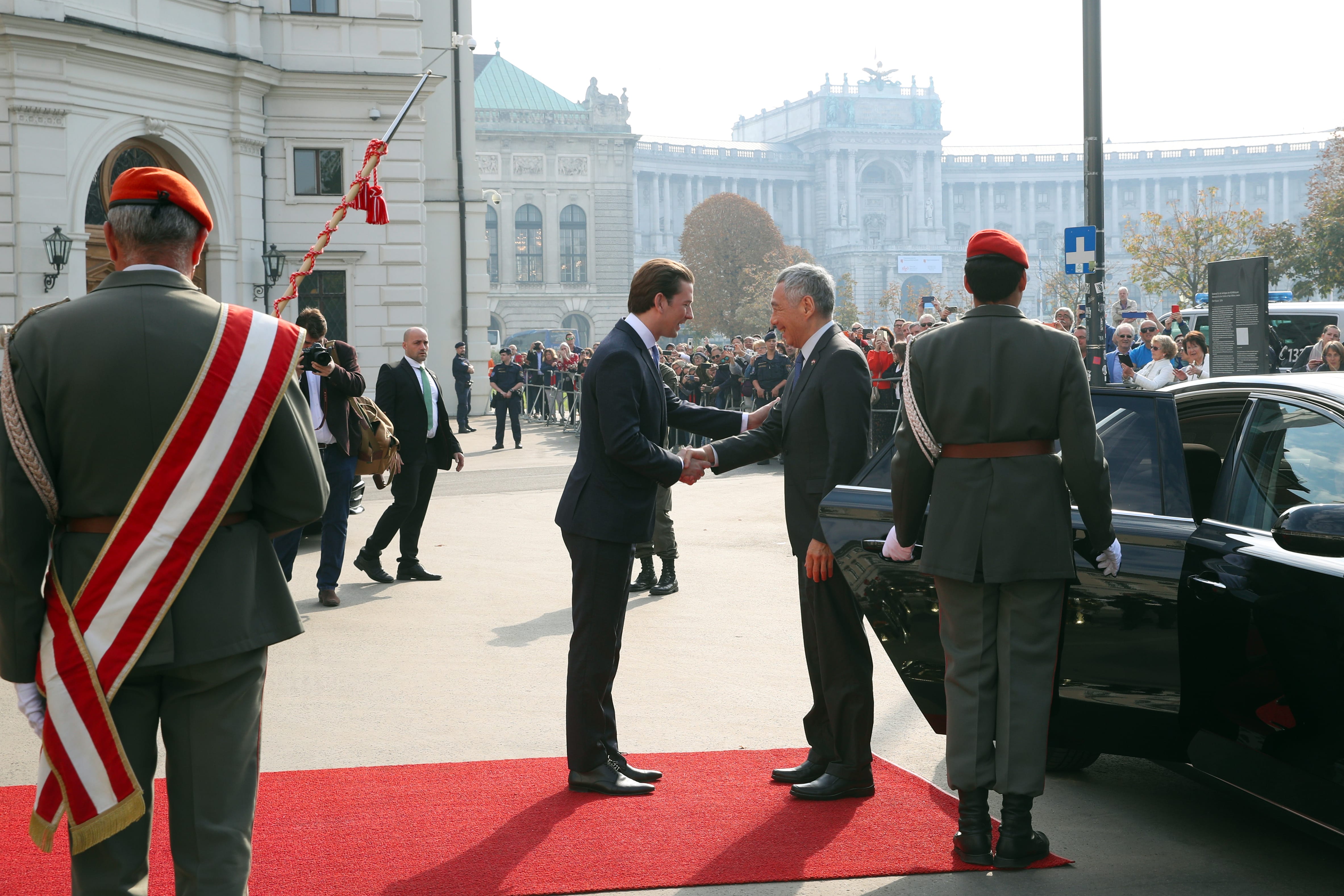 Two men in suits shake hands on a red carpet, soldiers in the foreground, crowd and building behind.