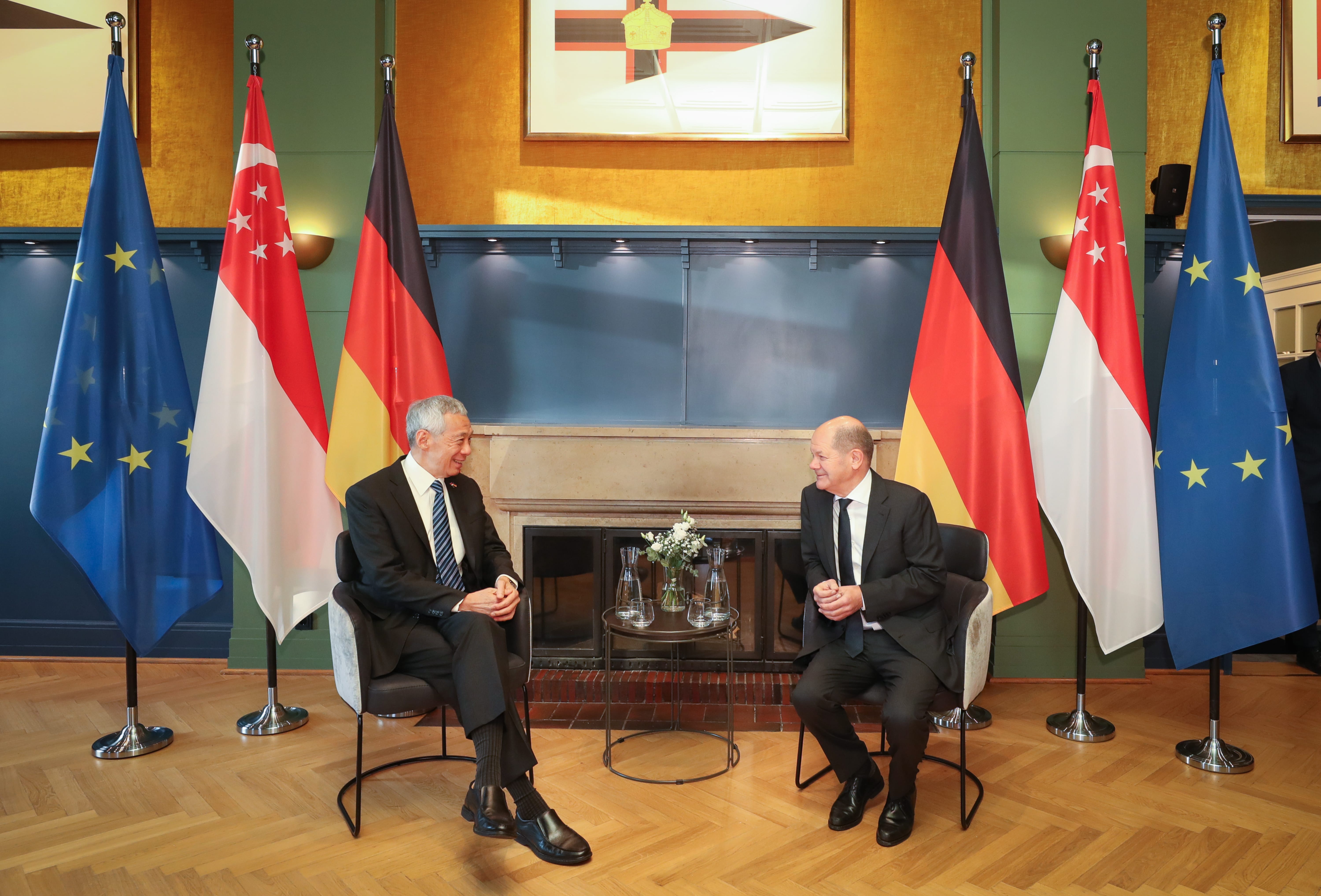 Lee Hsien Loong and Olaf Scholz sit in chairs with Singaporean, EU, and German flags behind them.
