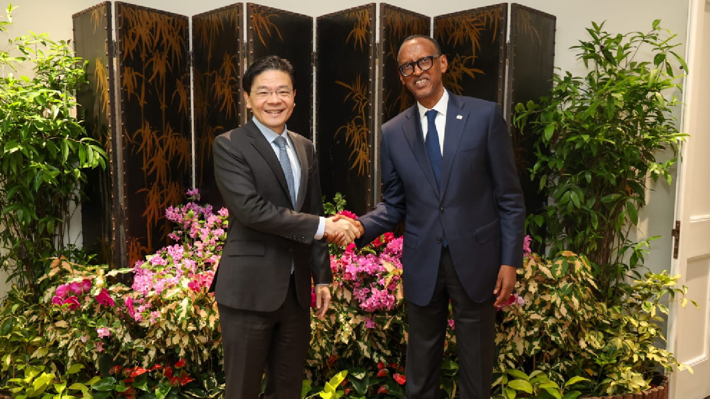 Two men in suits shaking hands in front of floral and foliage arrangements.