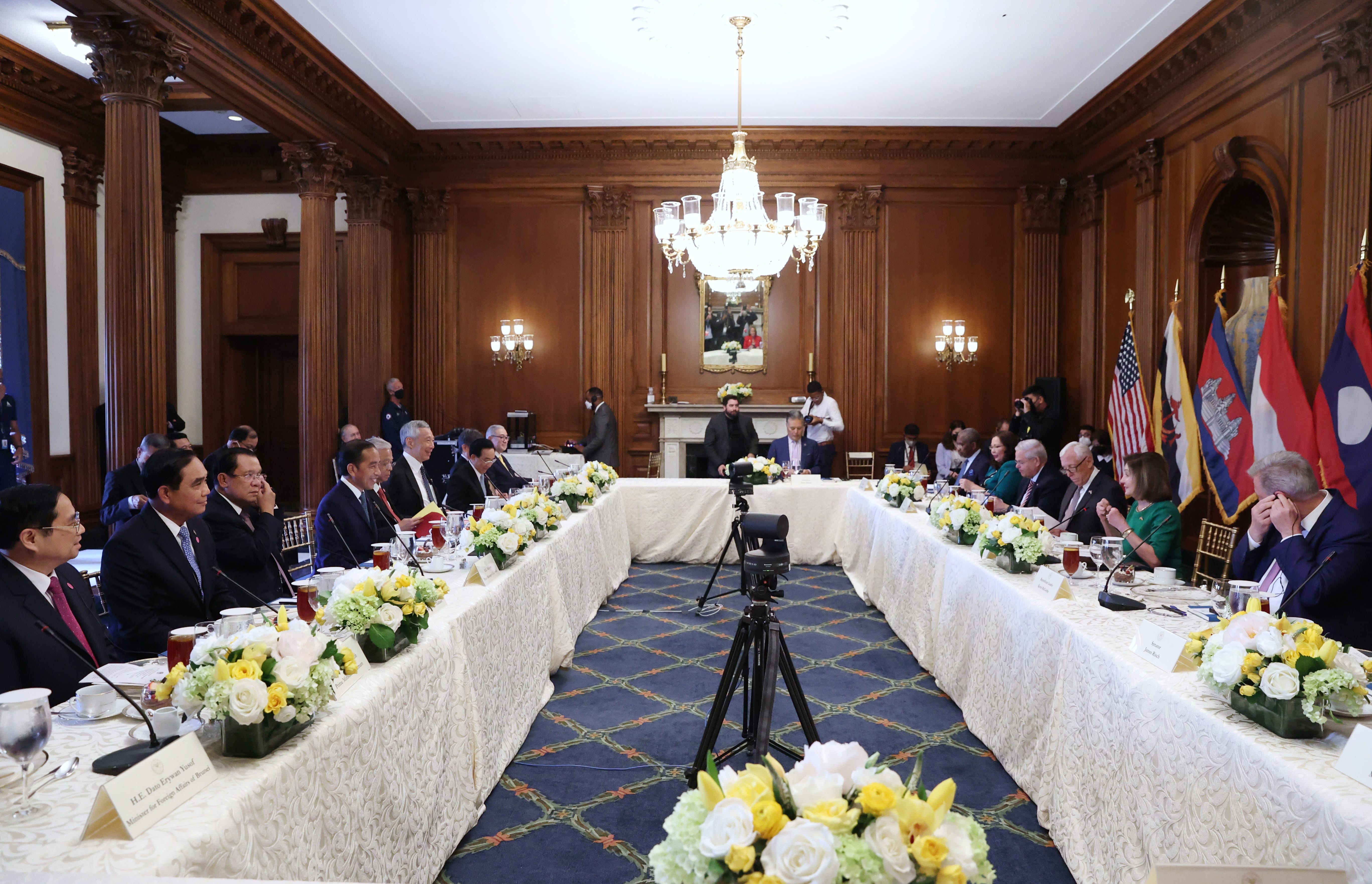 Long table meeting in wood-paneled room with flags, flowers, and camera tripod visible.