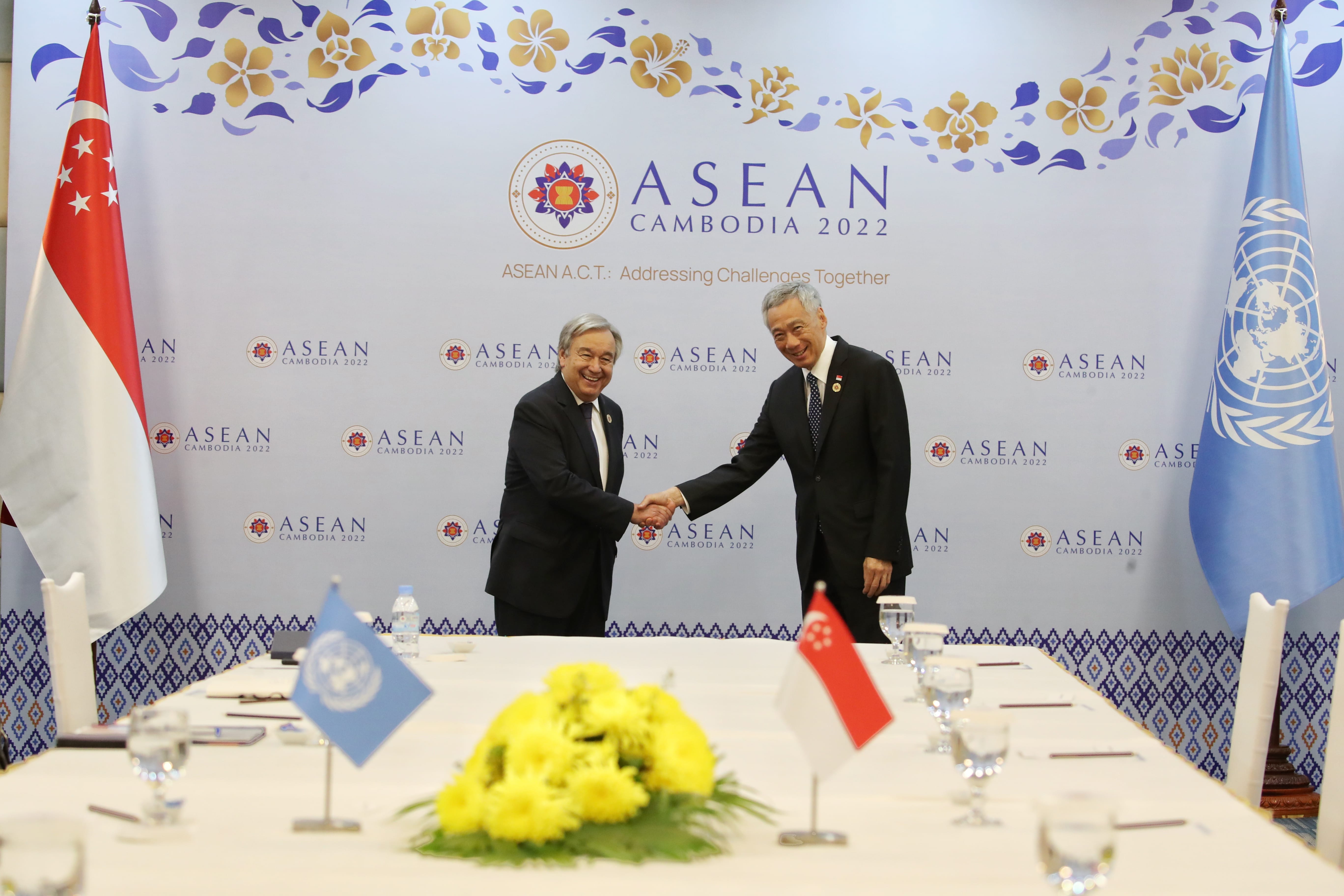 UN Secretary-General Guterres shakes hands with Singapore's Prime Minister Lee, flags on table, ASEAN backdrop.