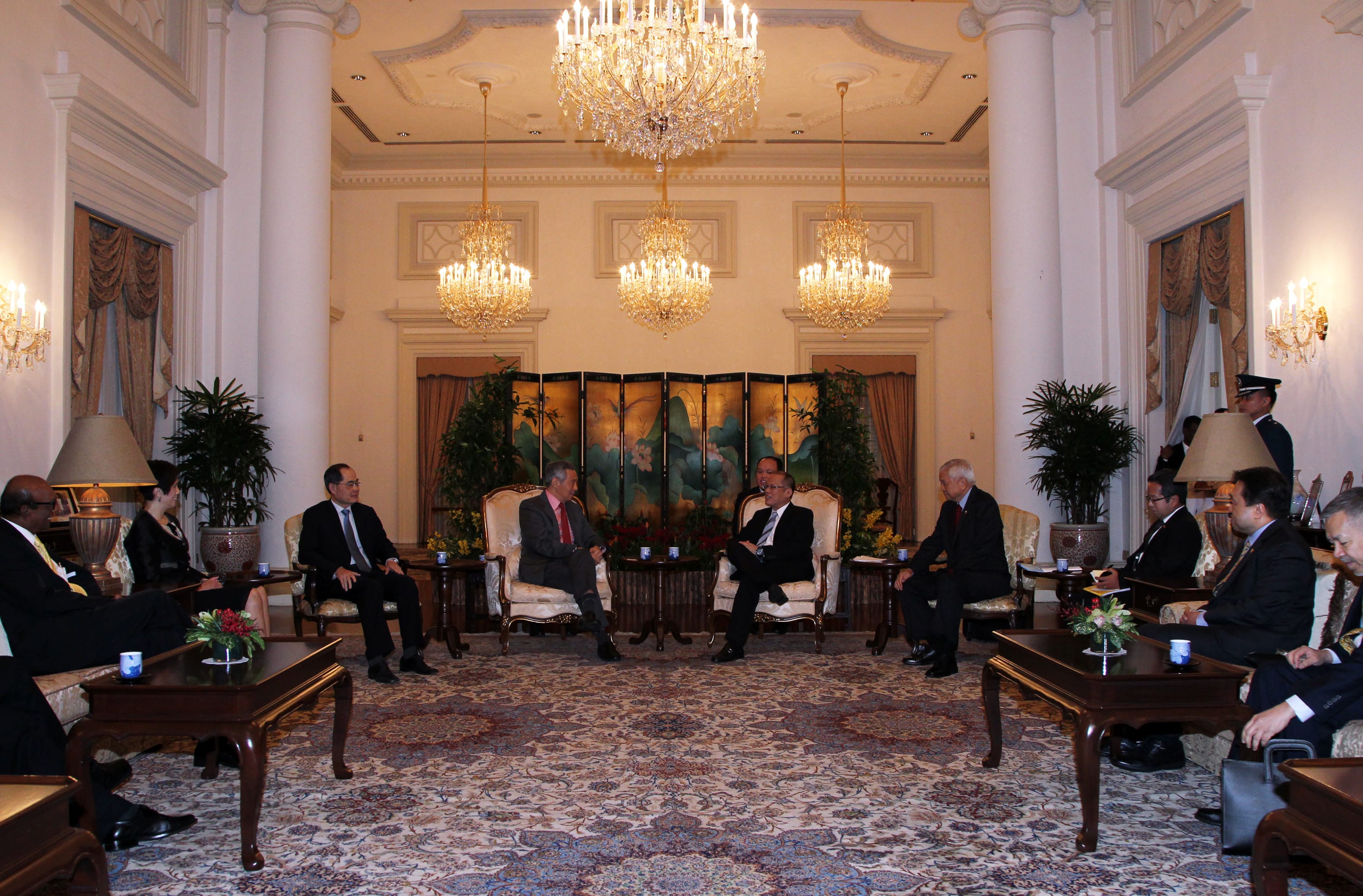 People seated in ornate room with columns, chandeliers, patterned rug, and dark wooden tables.