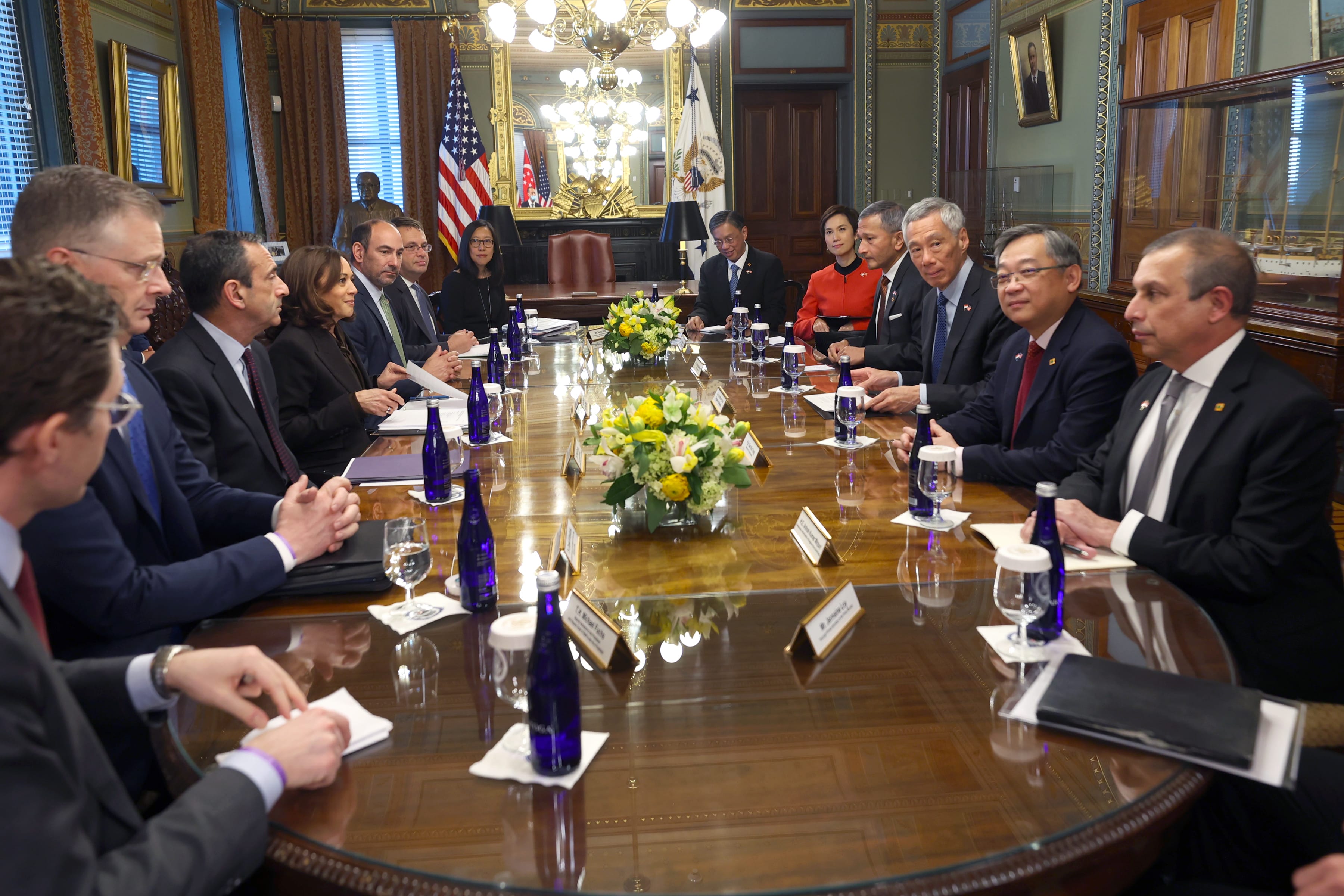 People sit around a conference table with flags, flowers, and water bottles. Kamala Harris is seated at the table.