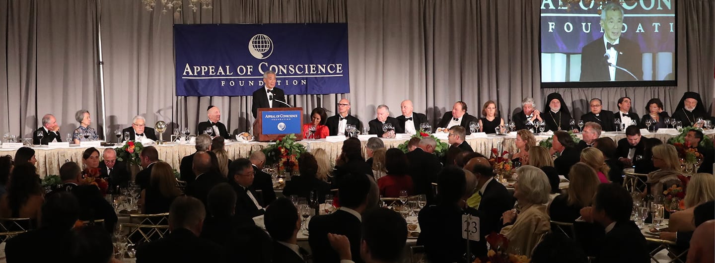 Banquet hall with speaker at podium below "Appeal of Conscience Foundation" banner, screen displays a speaker.