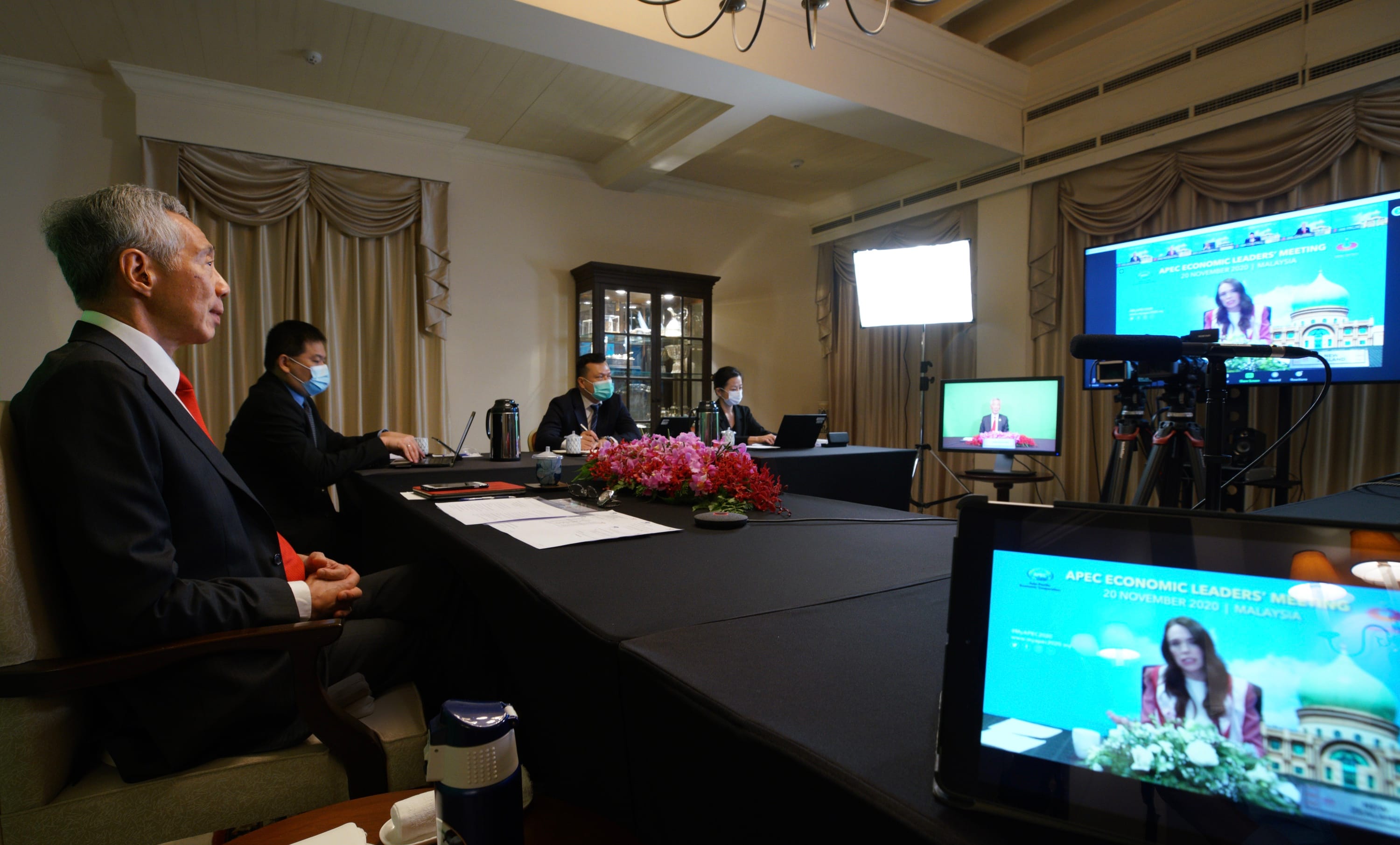 APEC meeting with attendees wearing masks, screens display leaders virtually.