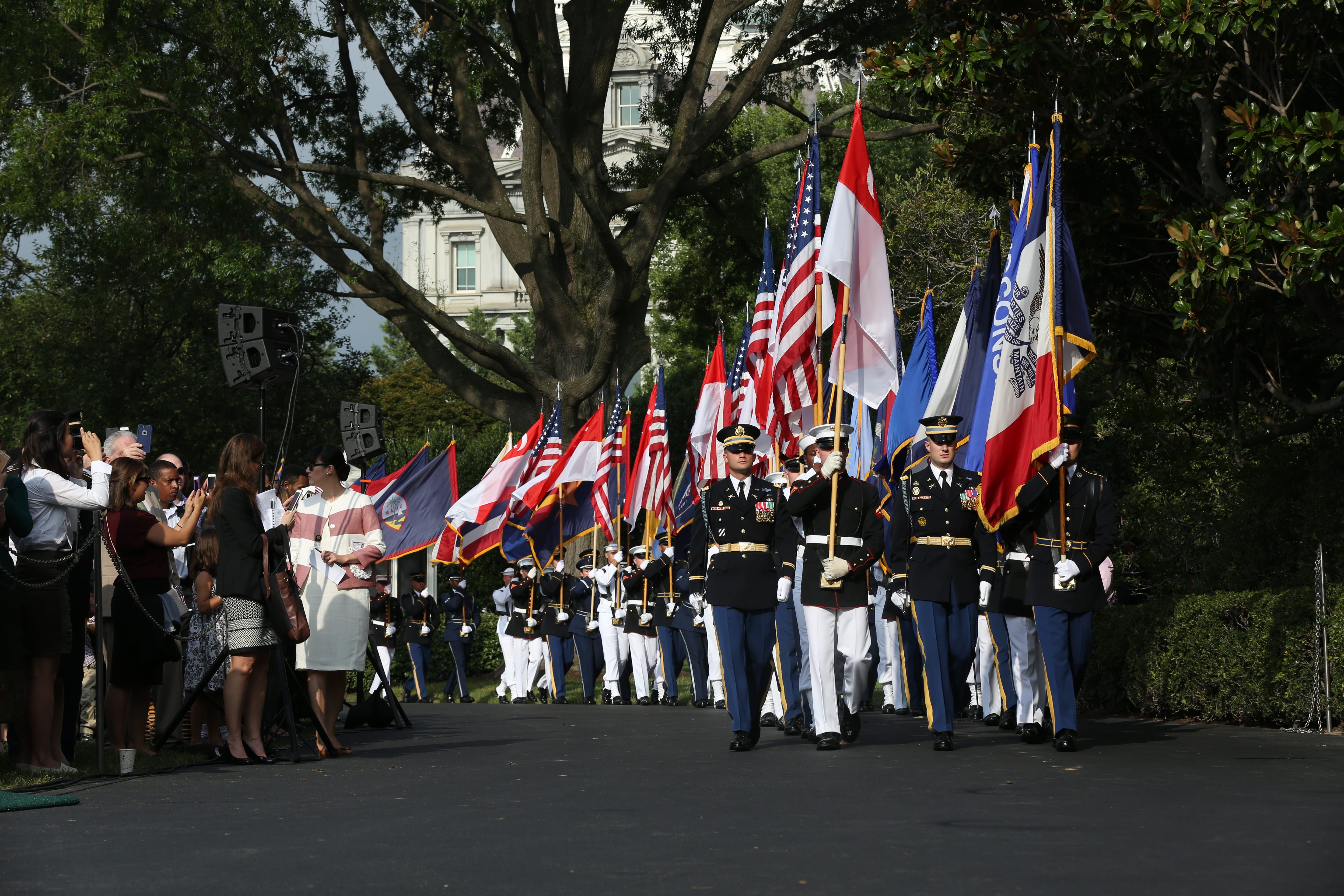 Military color guard in dress uniform marching with US flags and others; spectators taking photos.