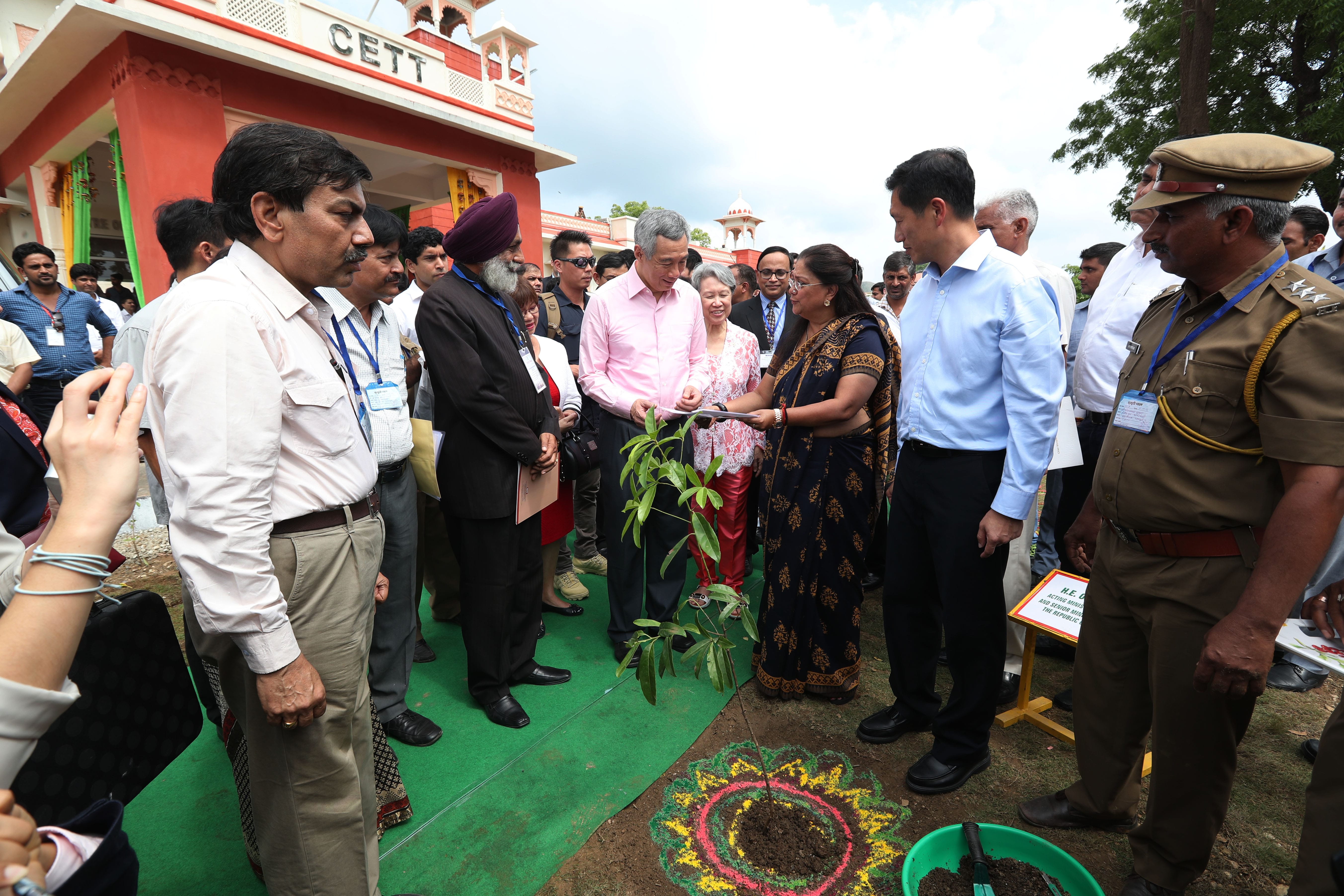 Group planting sapling in decorated soil outside CETT building, surrounded by onlookers.