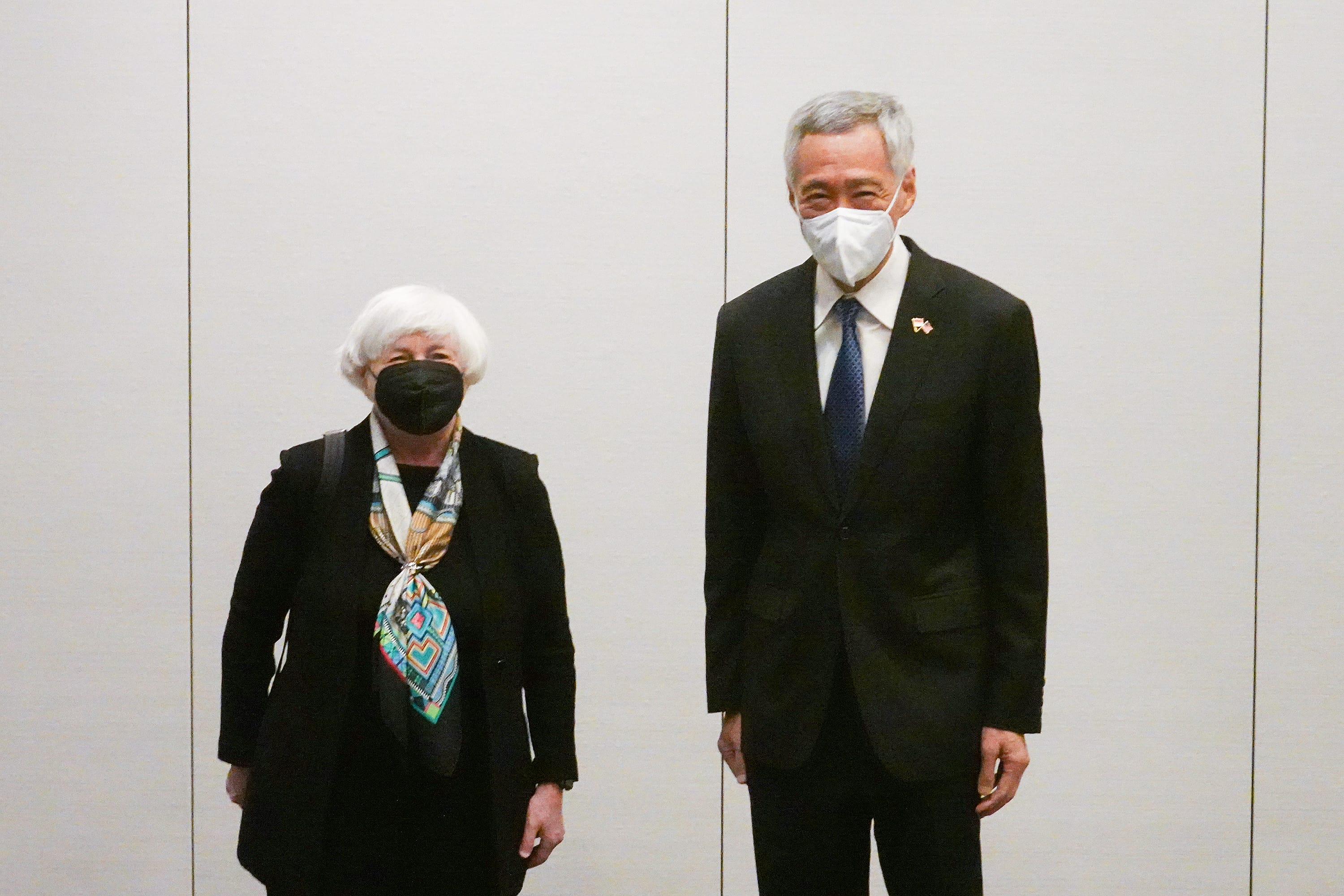 Janet Yellen & Lee Hsien Loong wear masks, dark suits, stand against a pale wall. Yellen wears a patterned scarf.