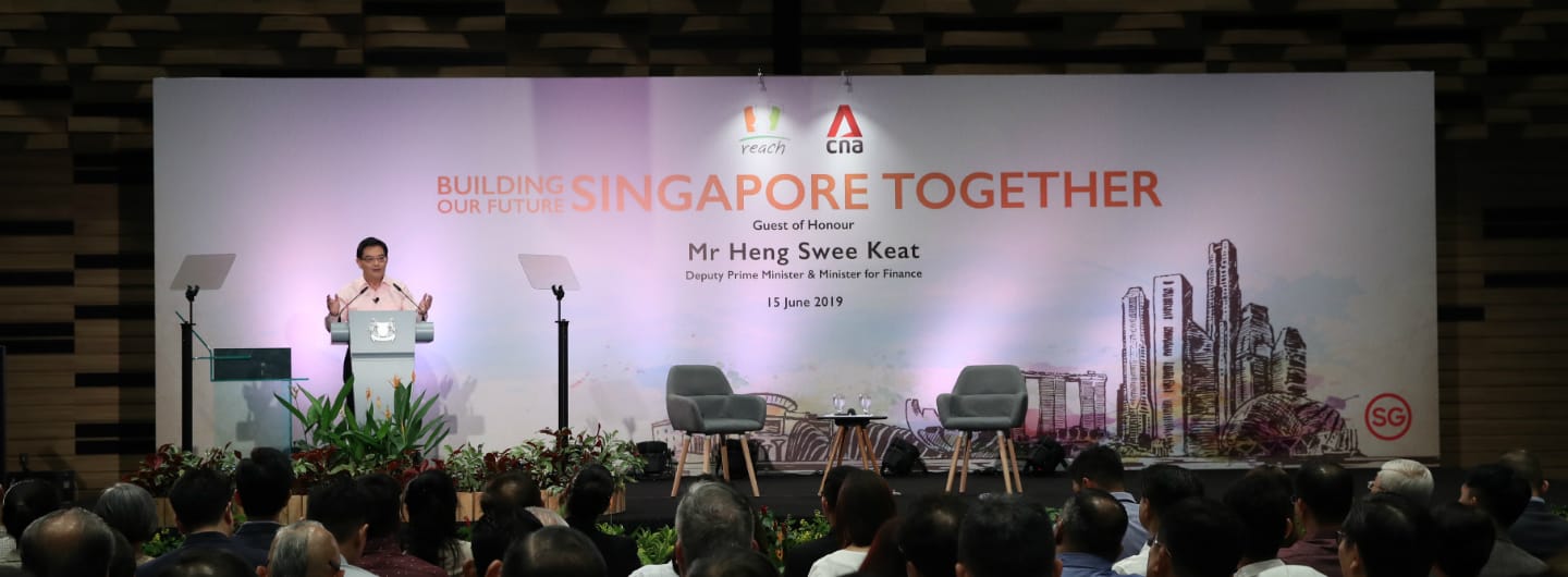 Man at podium with "Building Singapore Together" backdrop before audience, chairs, and Singapore skyline.