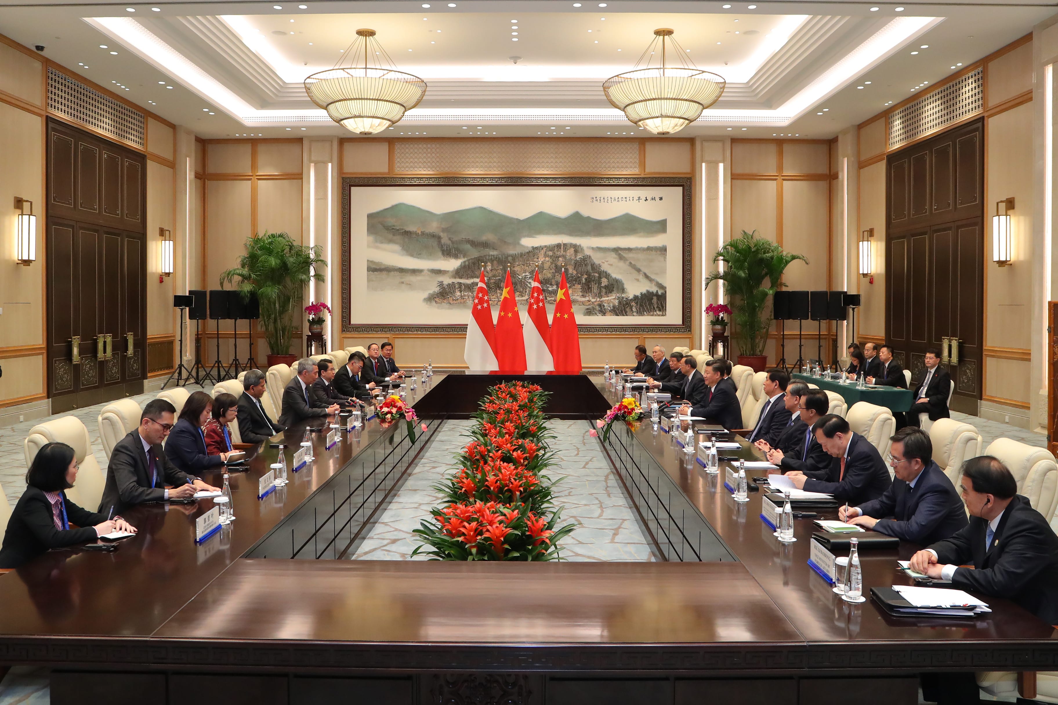 Formal meeting room with long table, seated attendees, China & Singapore flags, and floral centerpiece.