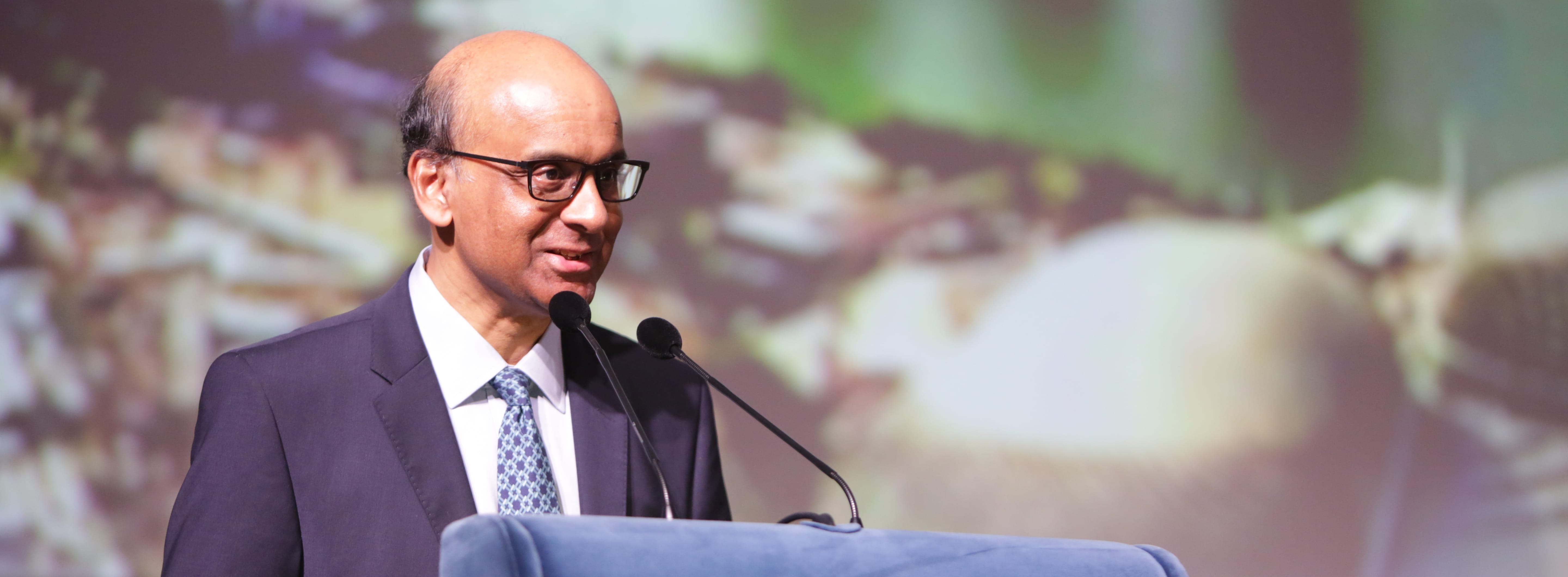 Man in suit with glasses speaking at podium with microphones; blurred background.