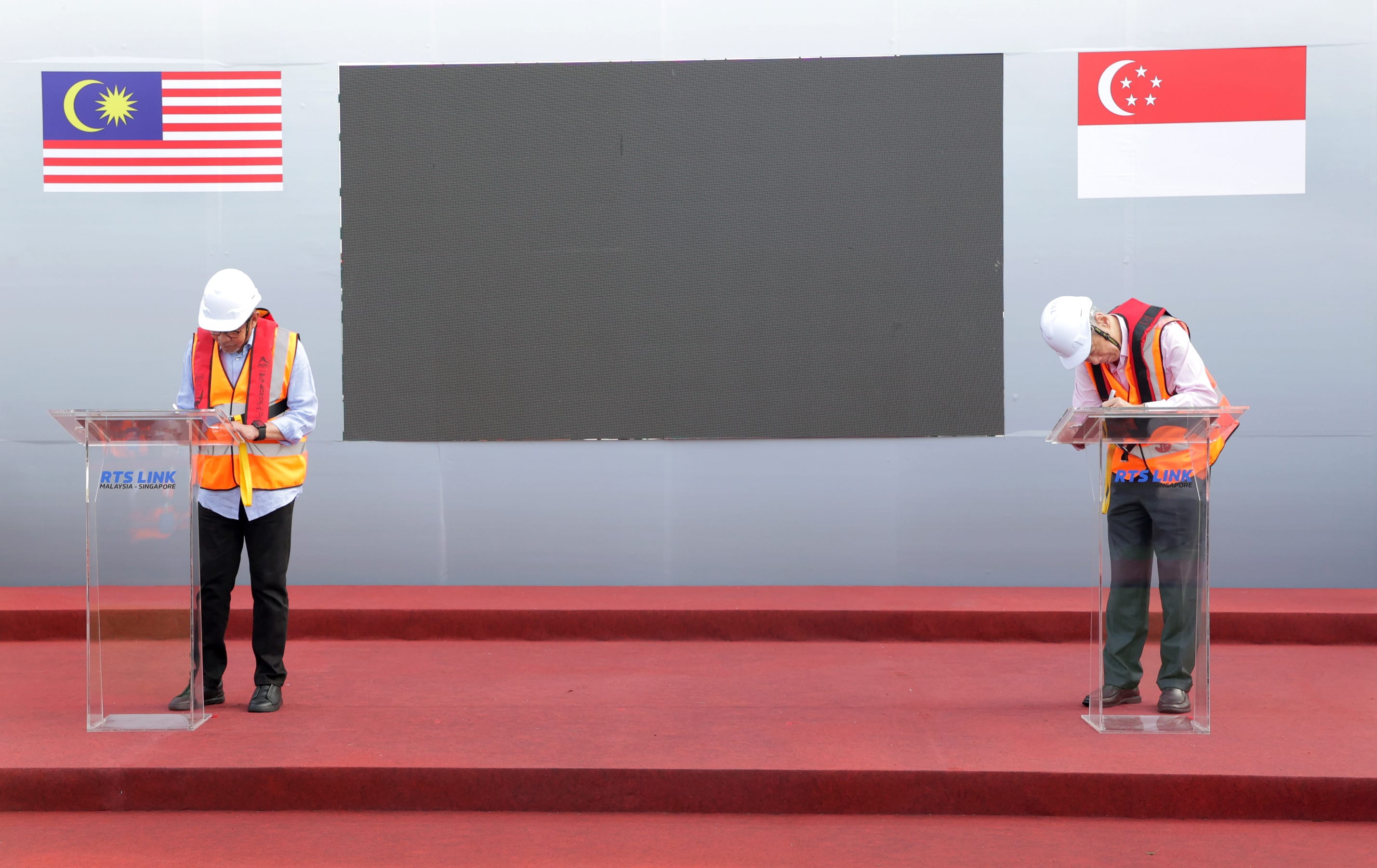 Two people sign RTS Link documents at podiums, flags of Malaysia and Singapore shown.