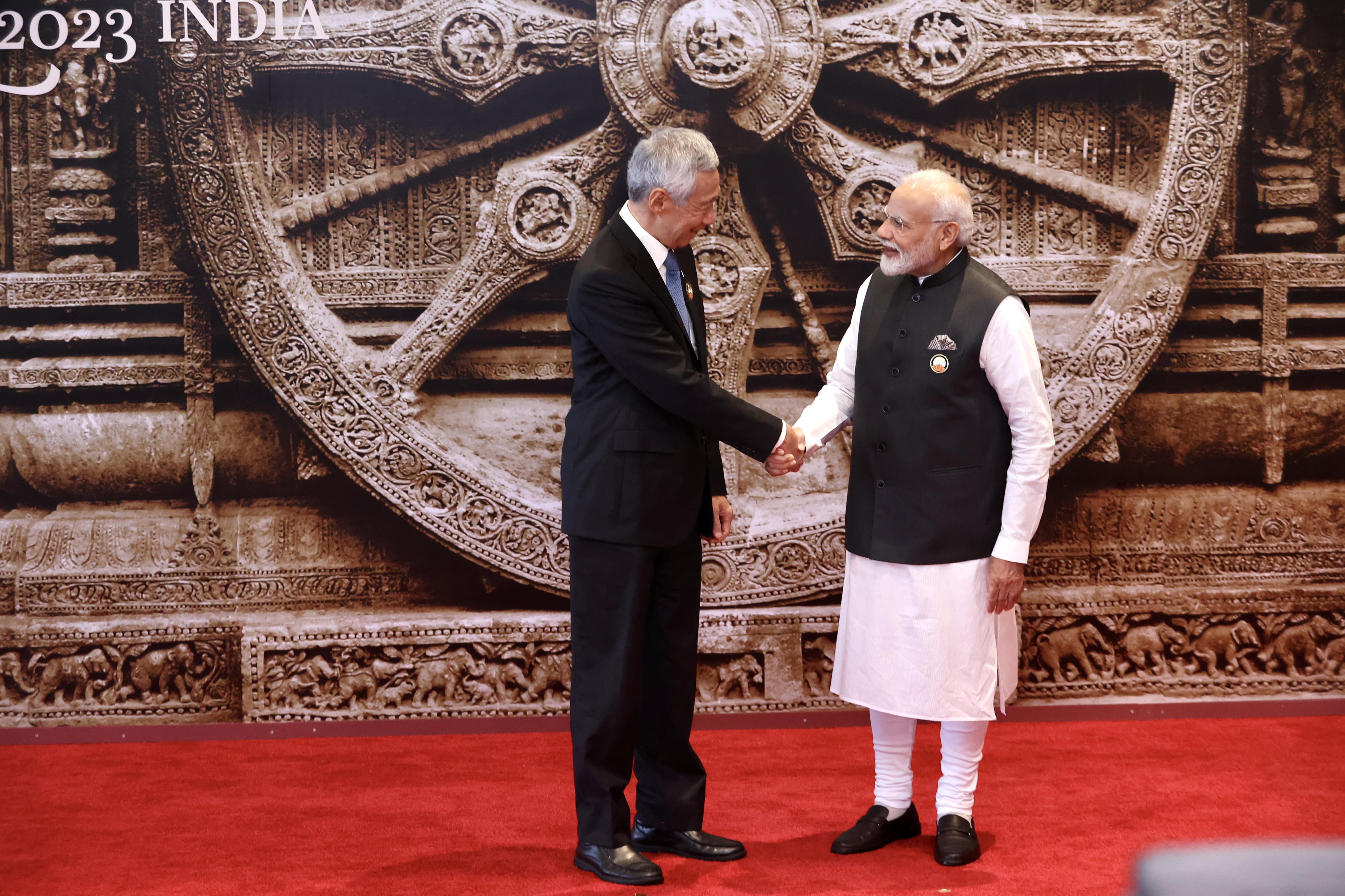 Two men shake hands in front of a backdrop with the Konark Wheel and "2023 INDIA".