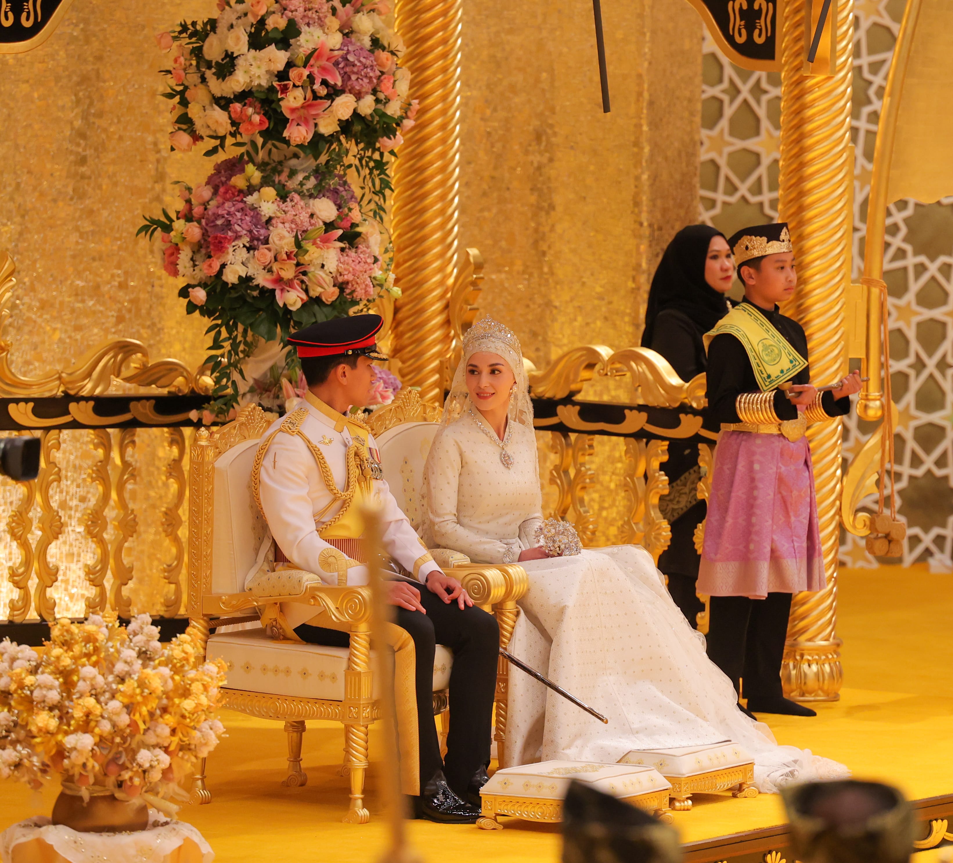 Brunei royal wedding: Bride in ornate white gown, groom in white military dress, on gold throne.