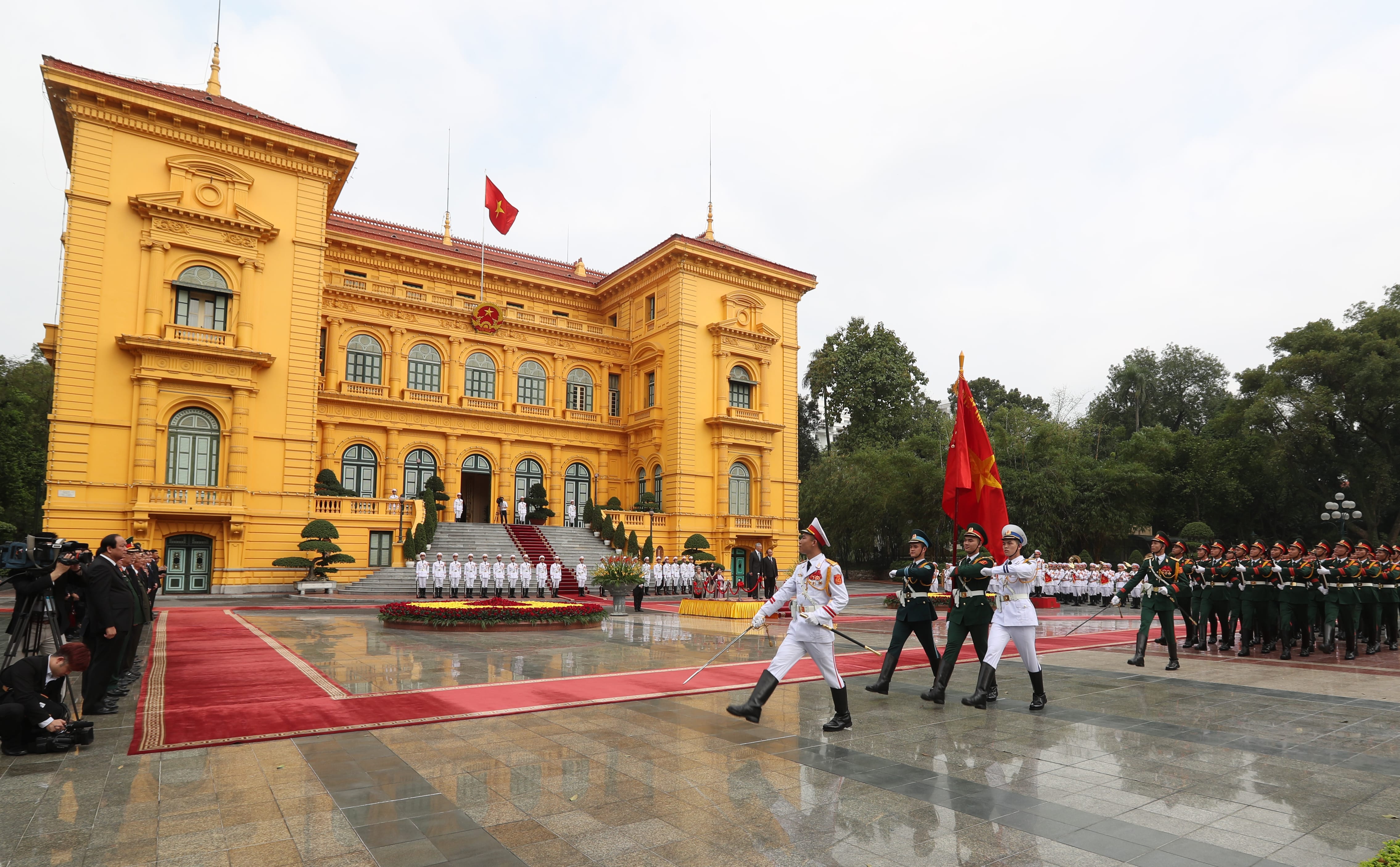 Military honor guard in formation before yellow palace, flag of Vietnam visible.