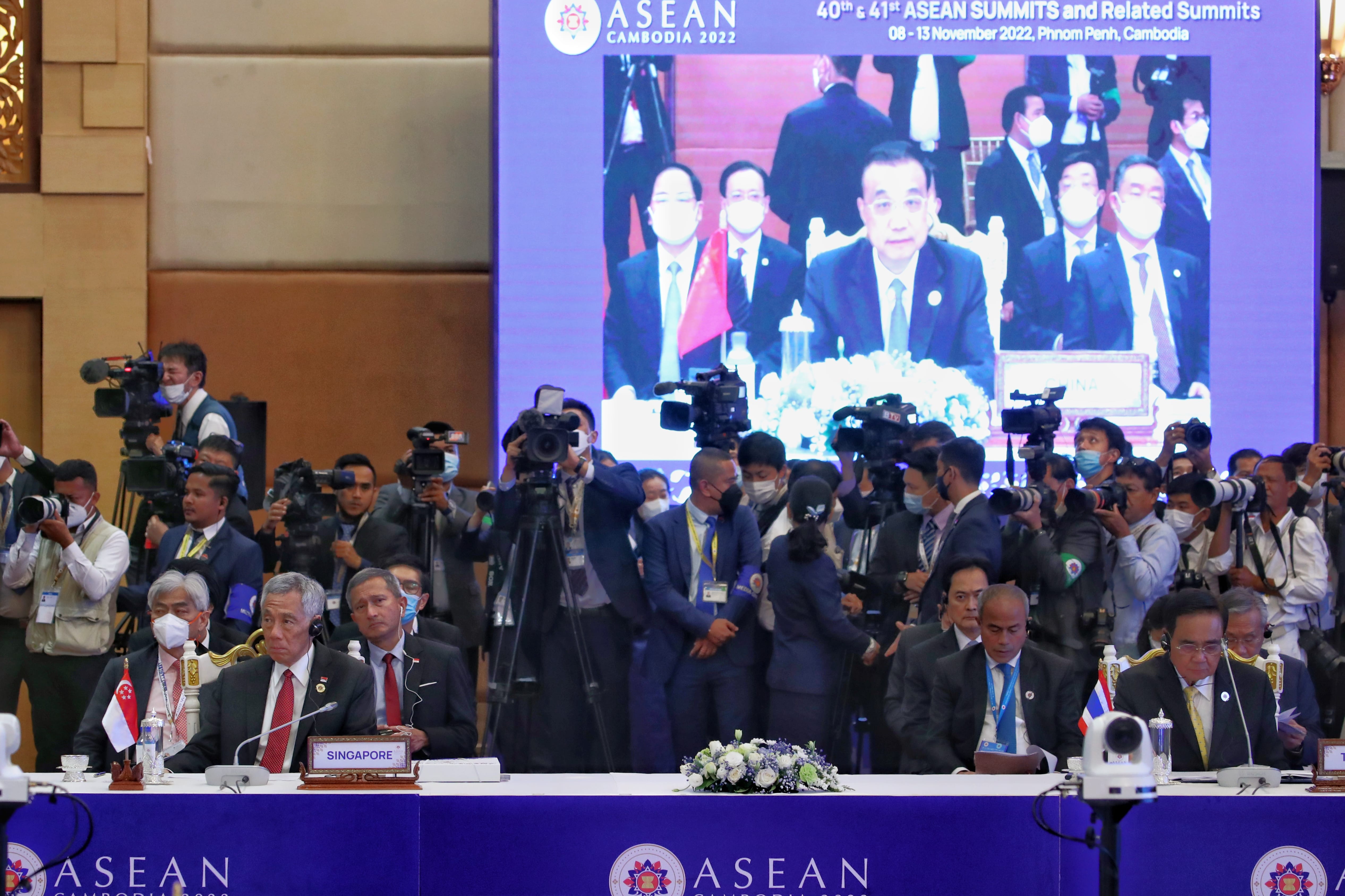 ASEAN summit with politicians at desks, cameras, and a large screen displaying people.