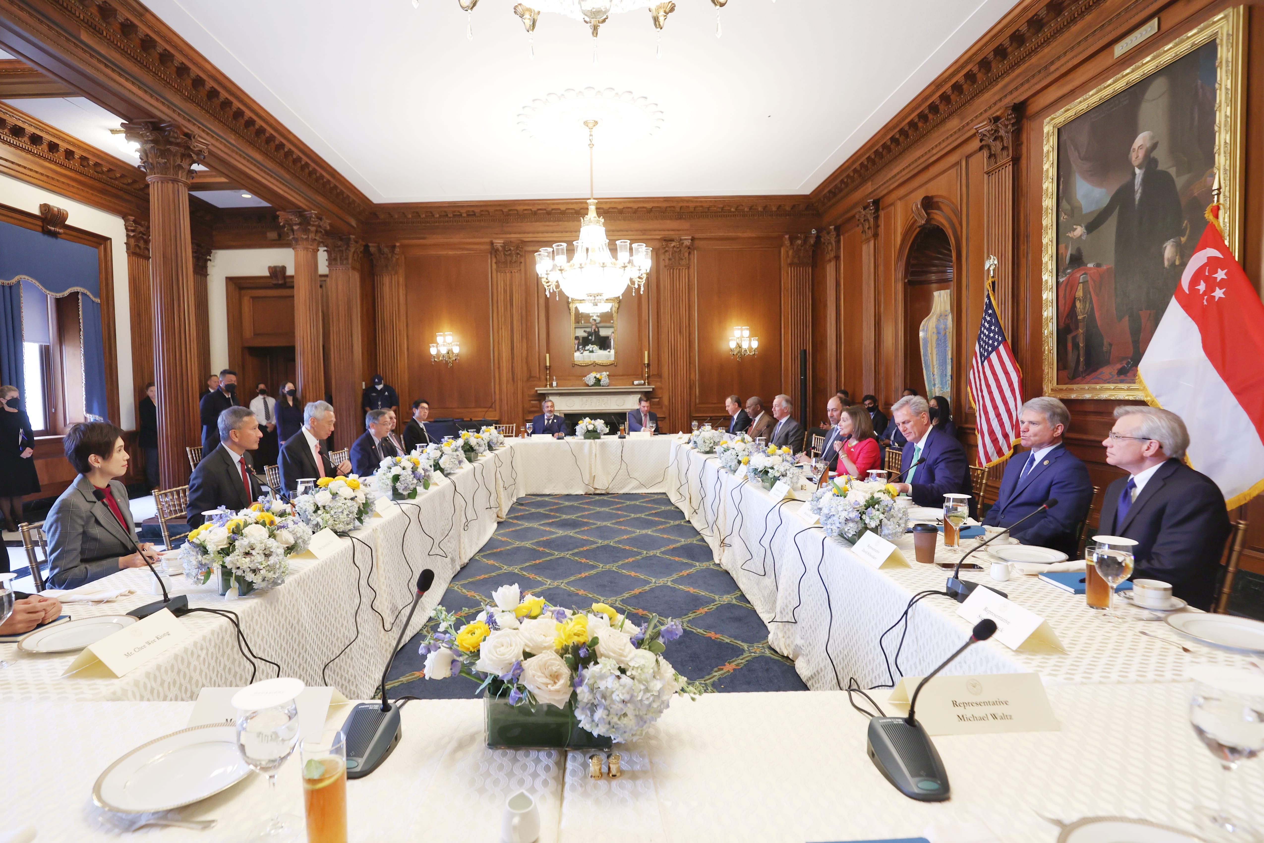 People at U-shaped table with microphones, flower arrangements, American and Singaporean flags. Wood-paneled room.