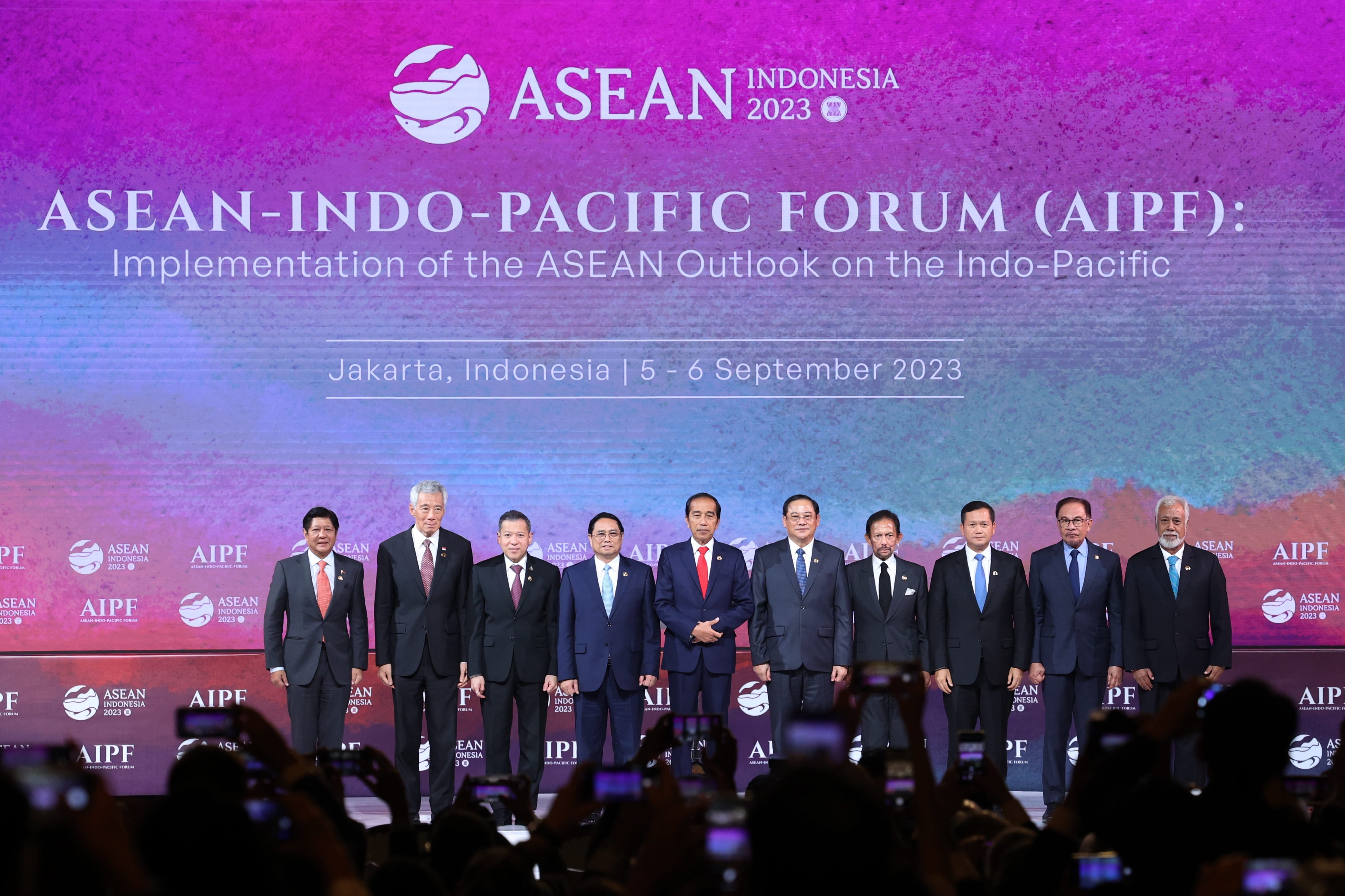 Ten people in suits stand on stage at the ASEAN-Indo-Pacific Forum (AIPF). Many hands hold up phones to photograph them.
