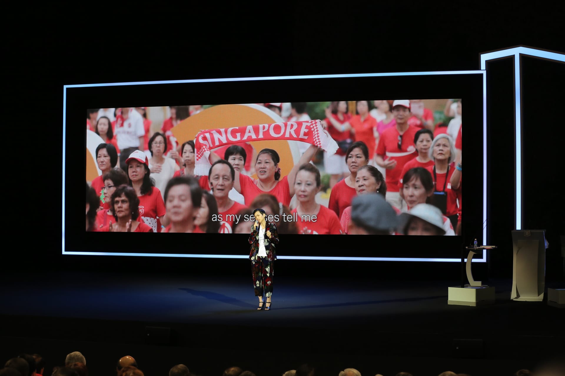 Woman on stage presents to a crowd; screen shows a crowd of people with a "Singapore" banner.