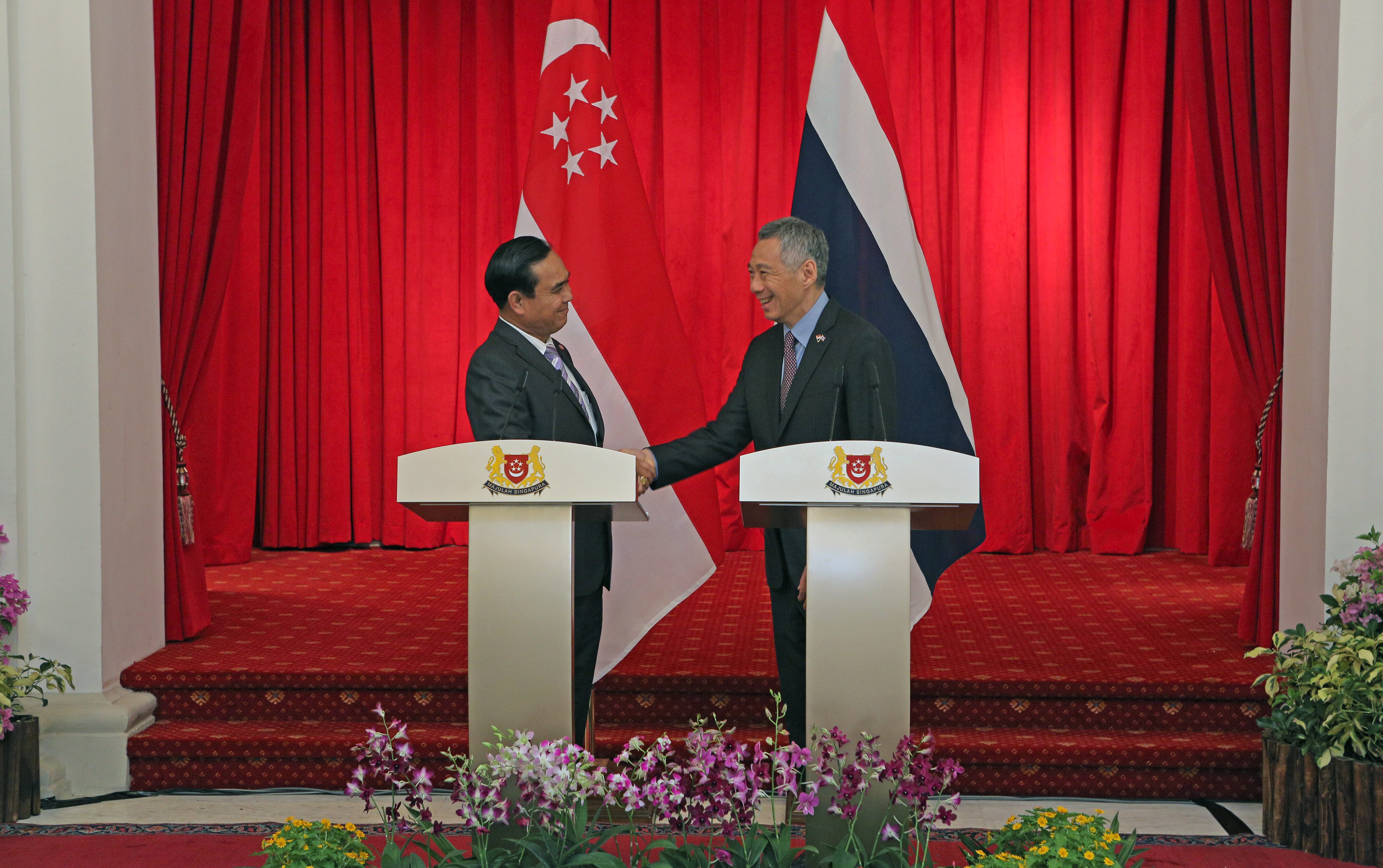 Two men shake hands at podiums displaying Singapore's crest, with Singapore and Thailand flags in the background.