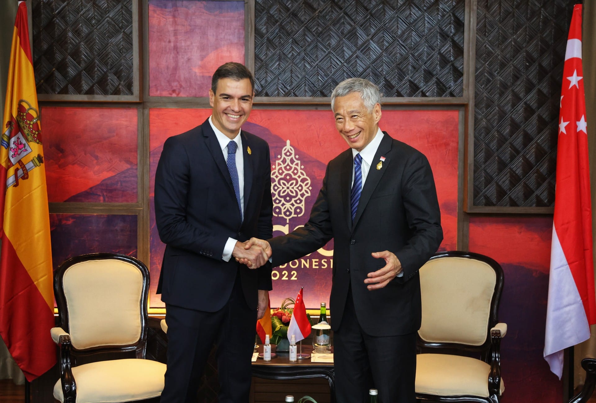Two men in suits shake hands, flanked by the flags of Spain and Singapore.