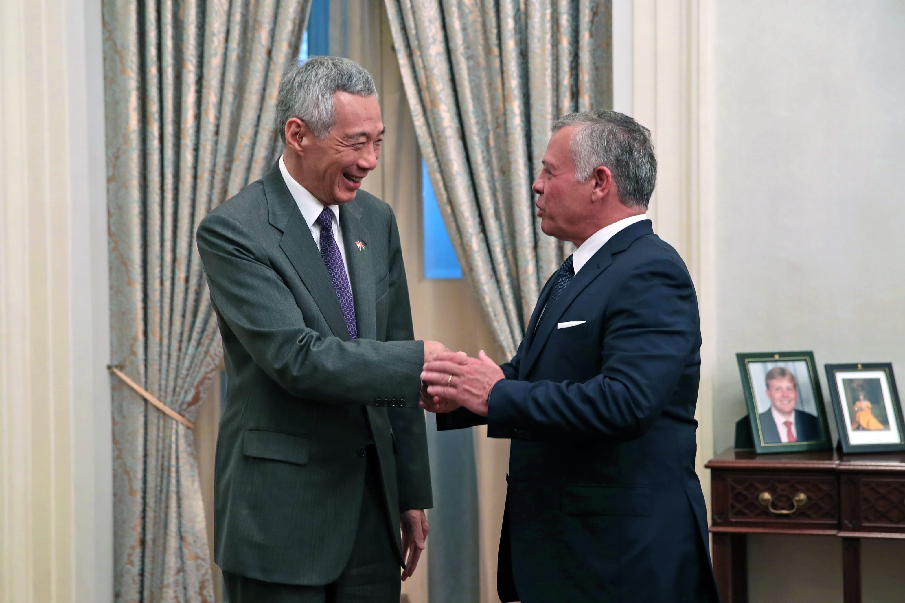 Two men in suits are shaking hands in front of patterned curtains.