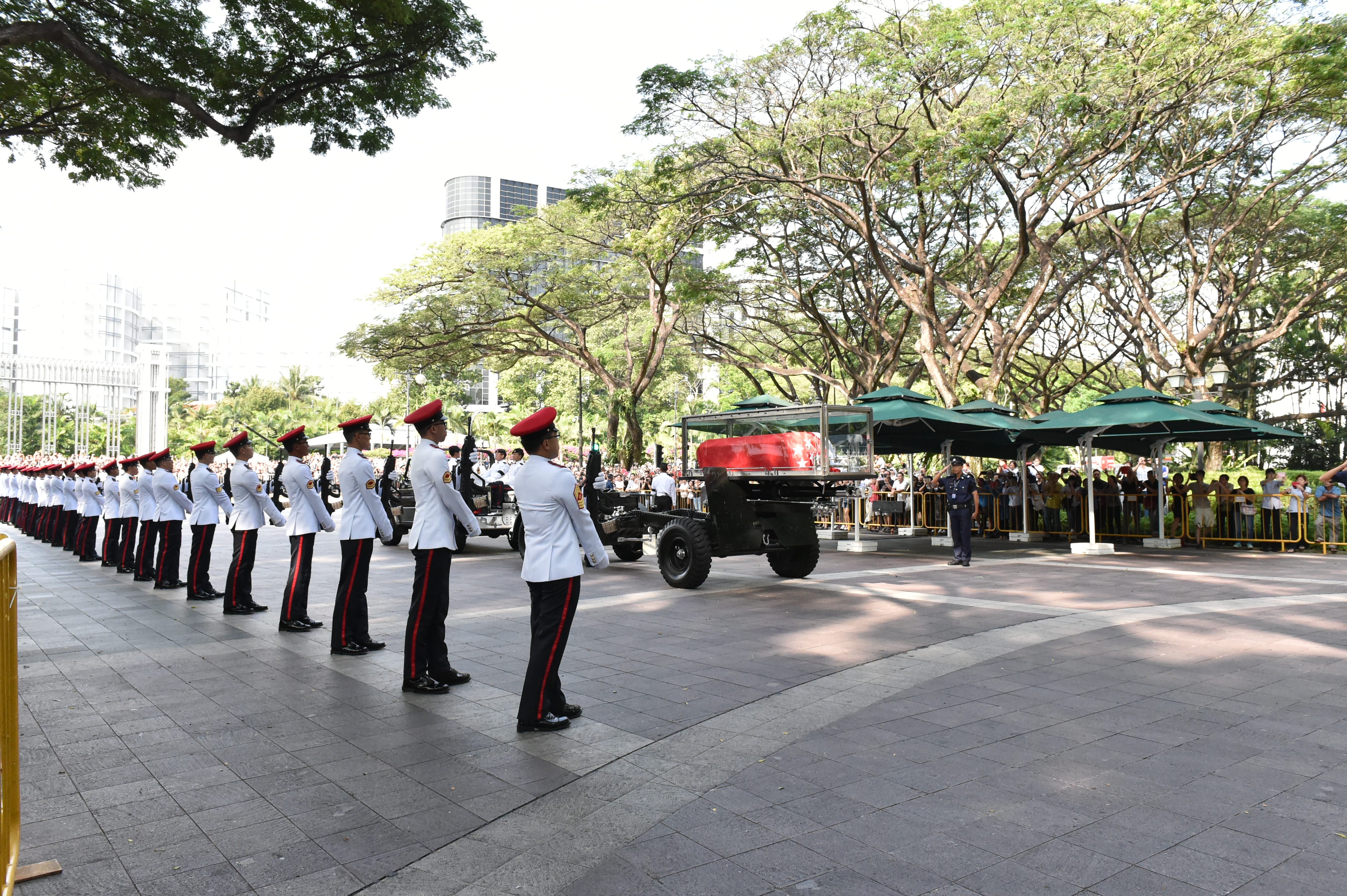 Ceremonial honor guard with rifles lines street beside hearse carrying coffin.