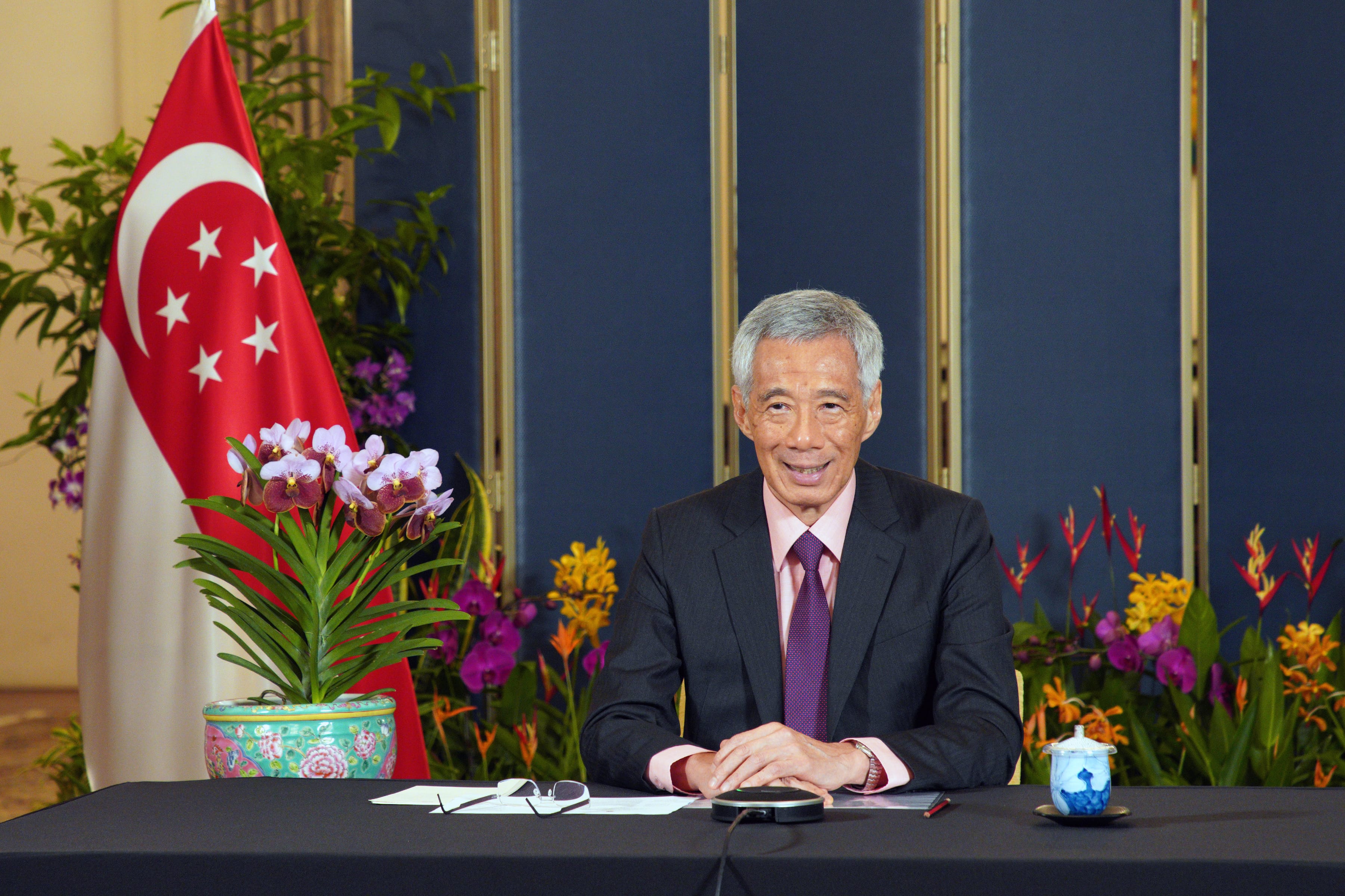 Lee Hsien Loong seated at a table with a Singapore flag and flowers in the background.