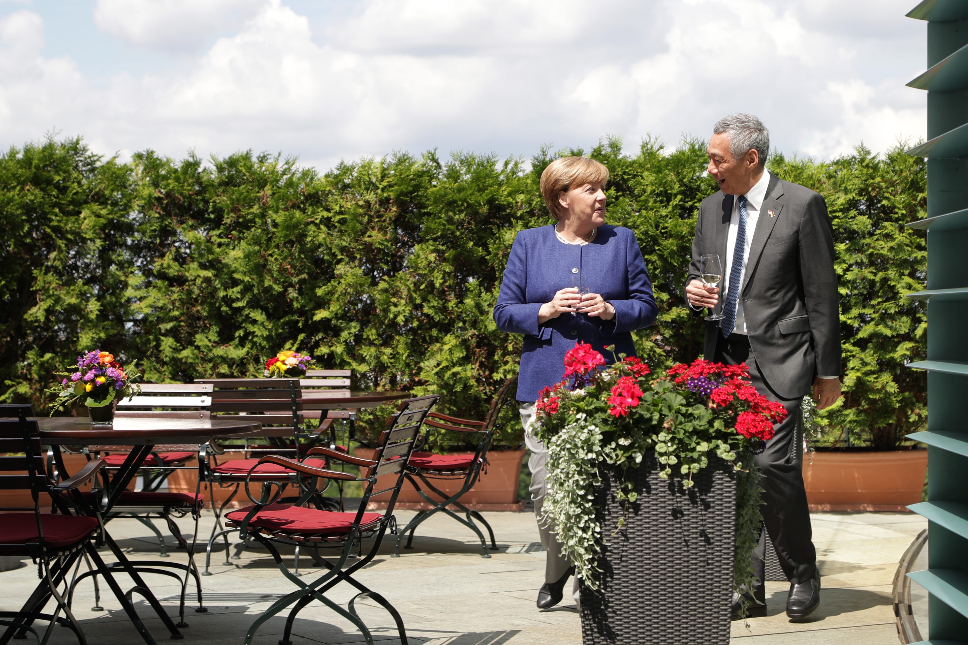 Angela Merkel & Lee Hsien Loong walk on patio holding wine glasses, with green hedges and outdoor seating behind them.