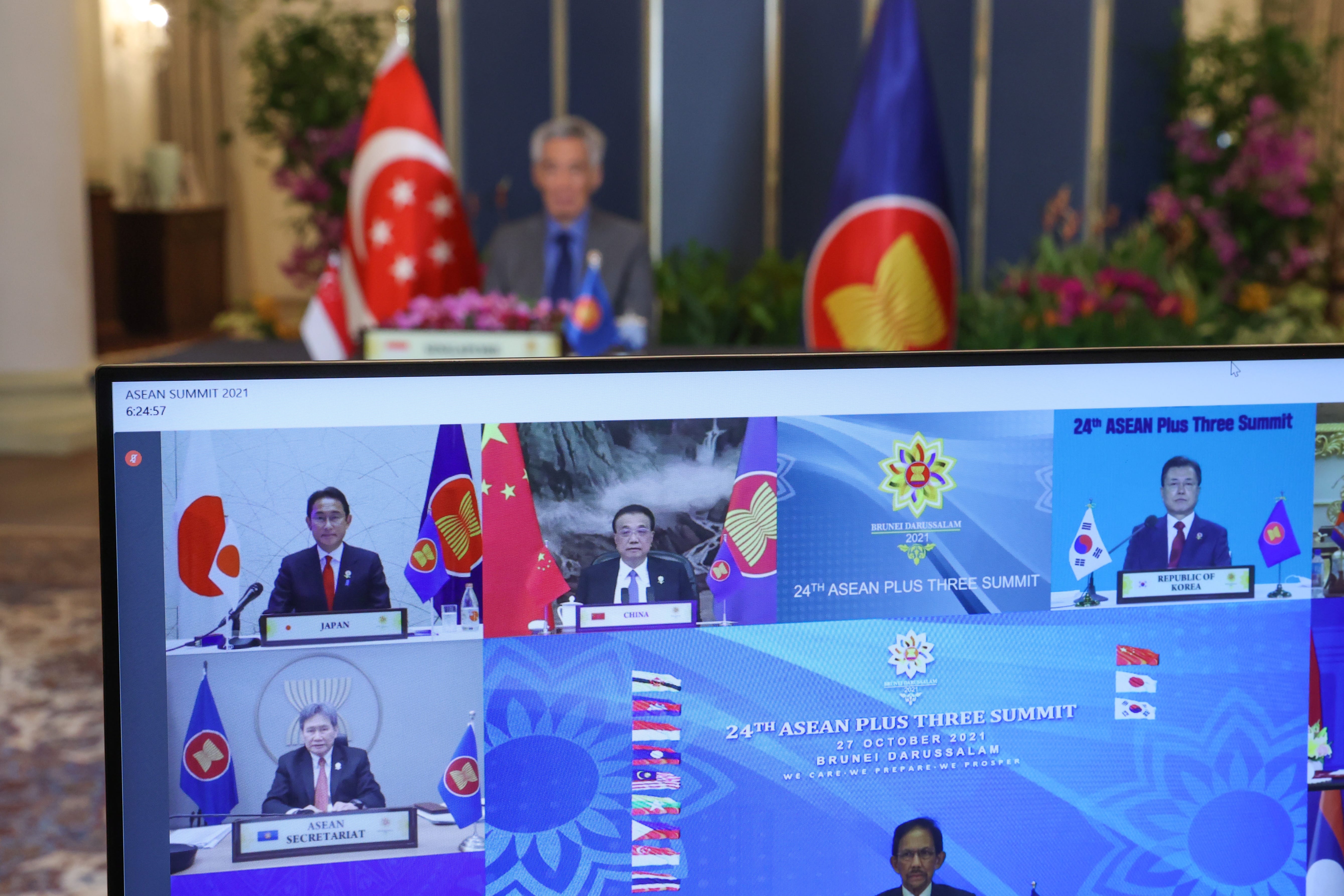 A monitor shows the ASEAN Summit 2021 virtual meeting with flags and leaders of Japan, China, and South Korea.