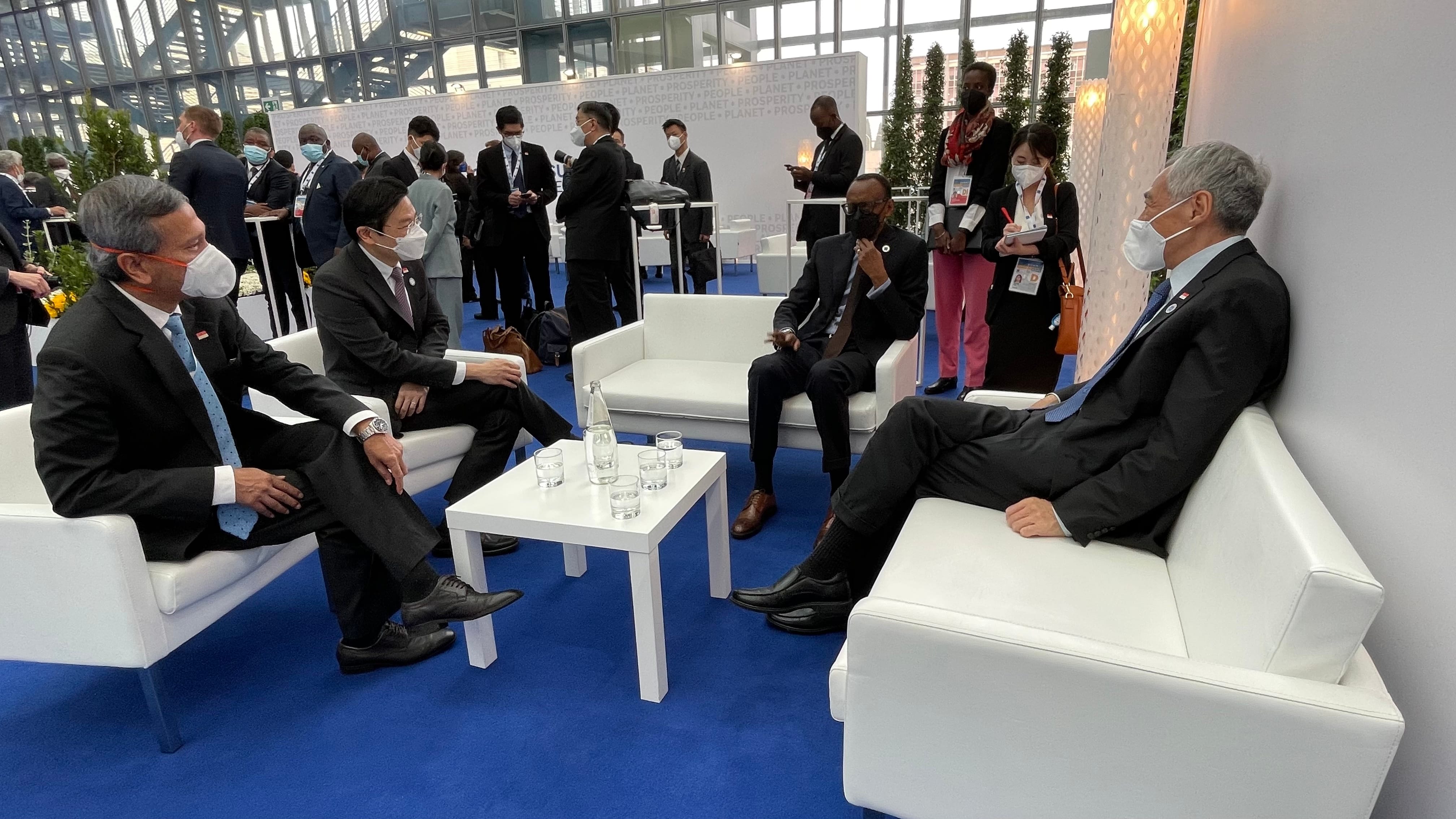 Group of people in suits with masks sit on white couches on blue carpet.
