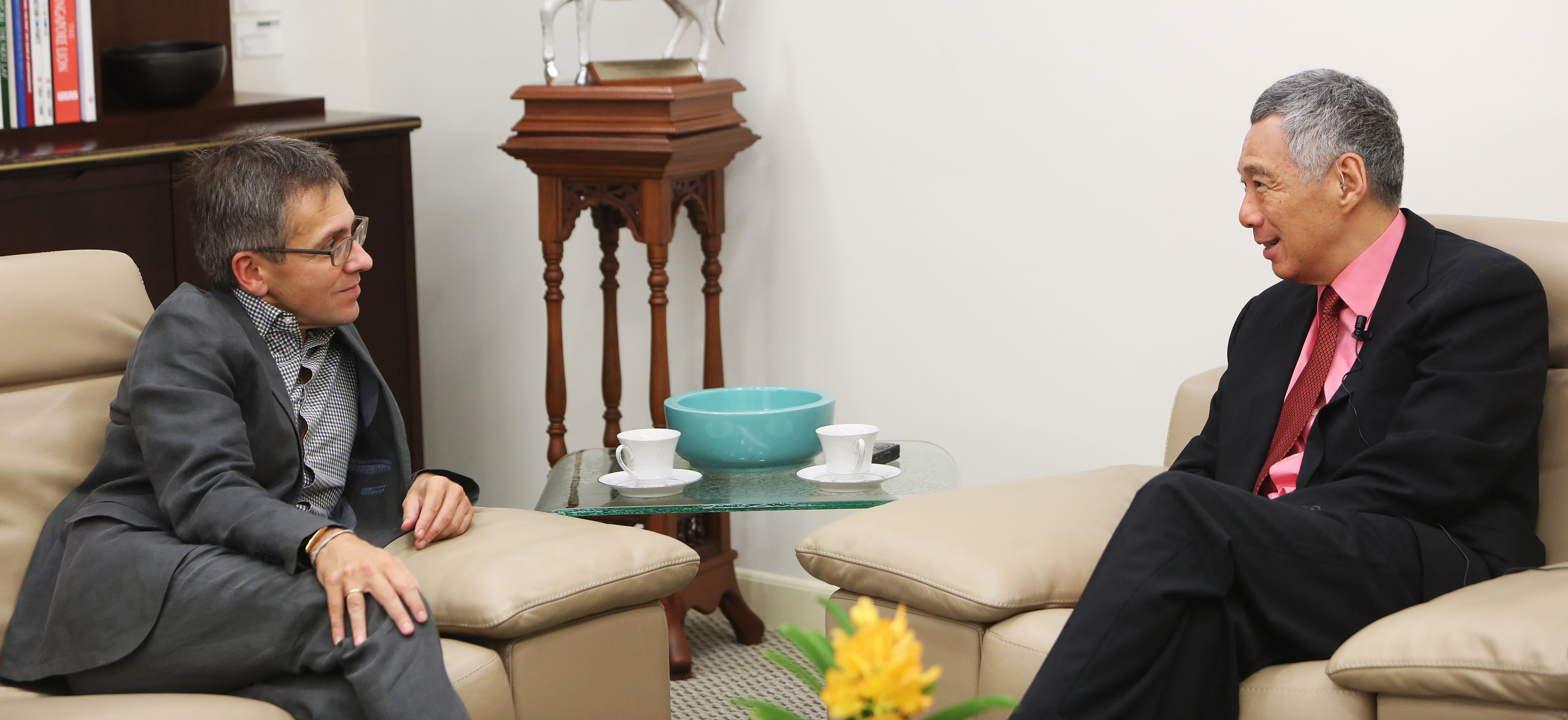 Two men in suits seated, facing each other, near a table with tea cups.