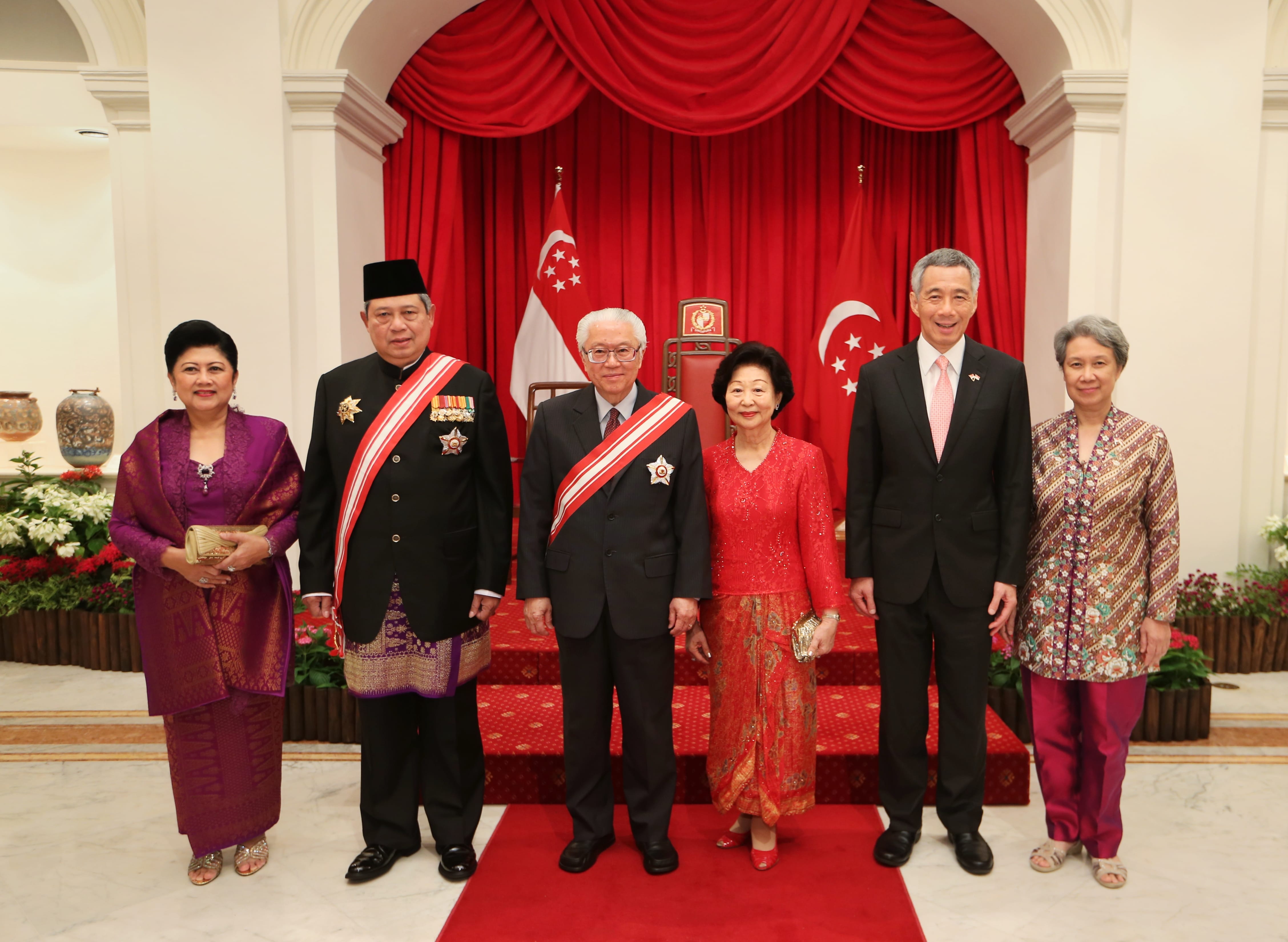 Six people pose before Singapore flags and red drapes in formal wear with sashes and medals.
