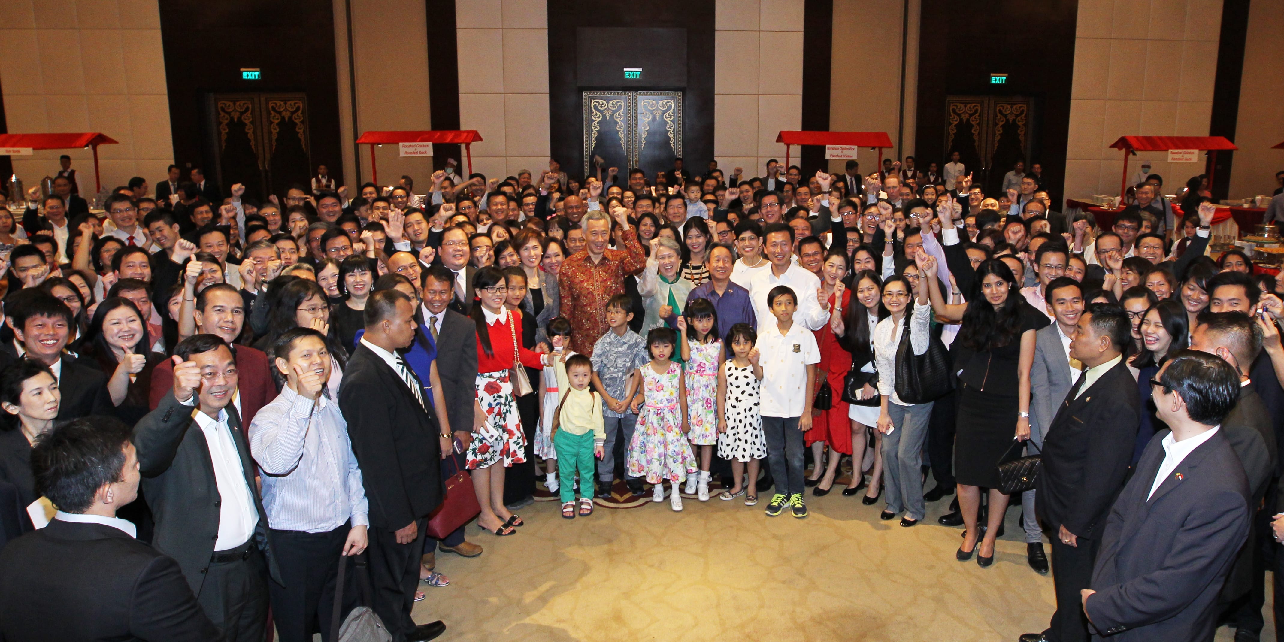 Large group poses, some giving thumbs-up, in a banquet hall with Lee Hsien Loong visible.