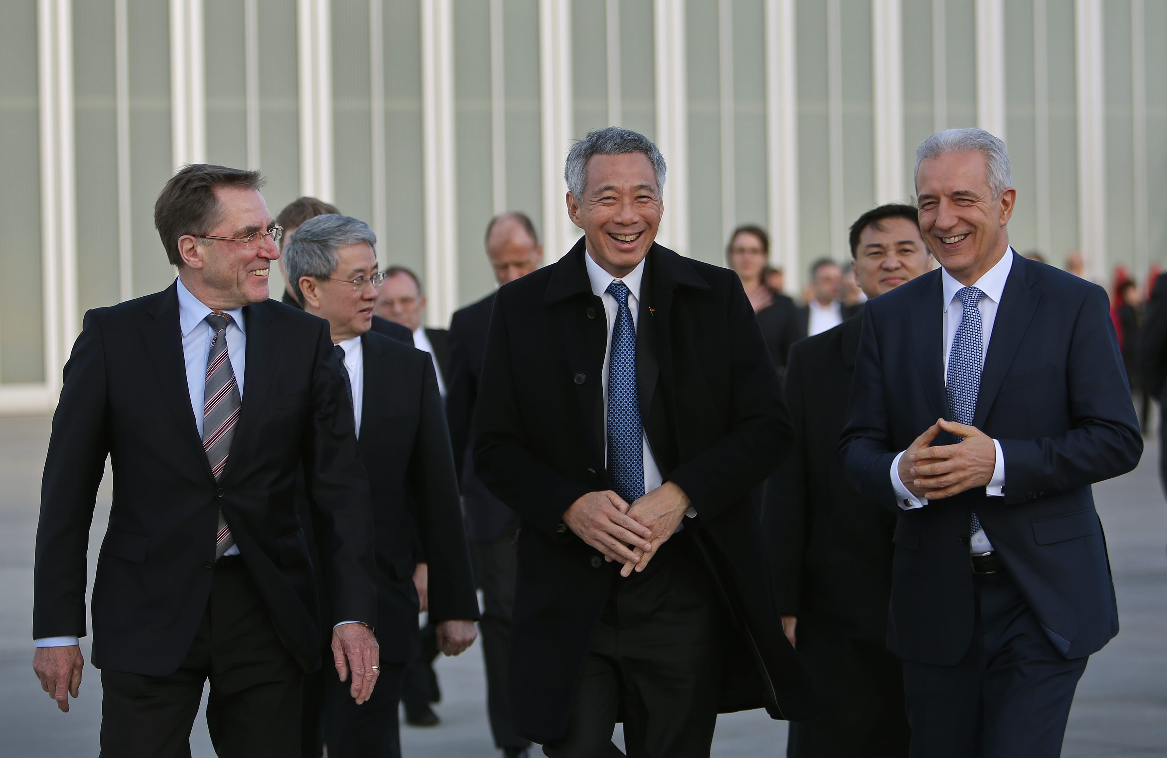 Group of men in suits, walking outside a building with vertical white panels. Lee Hsien Loong is in the center.