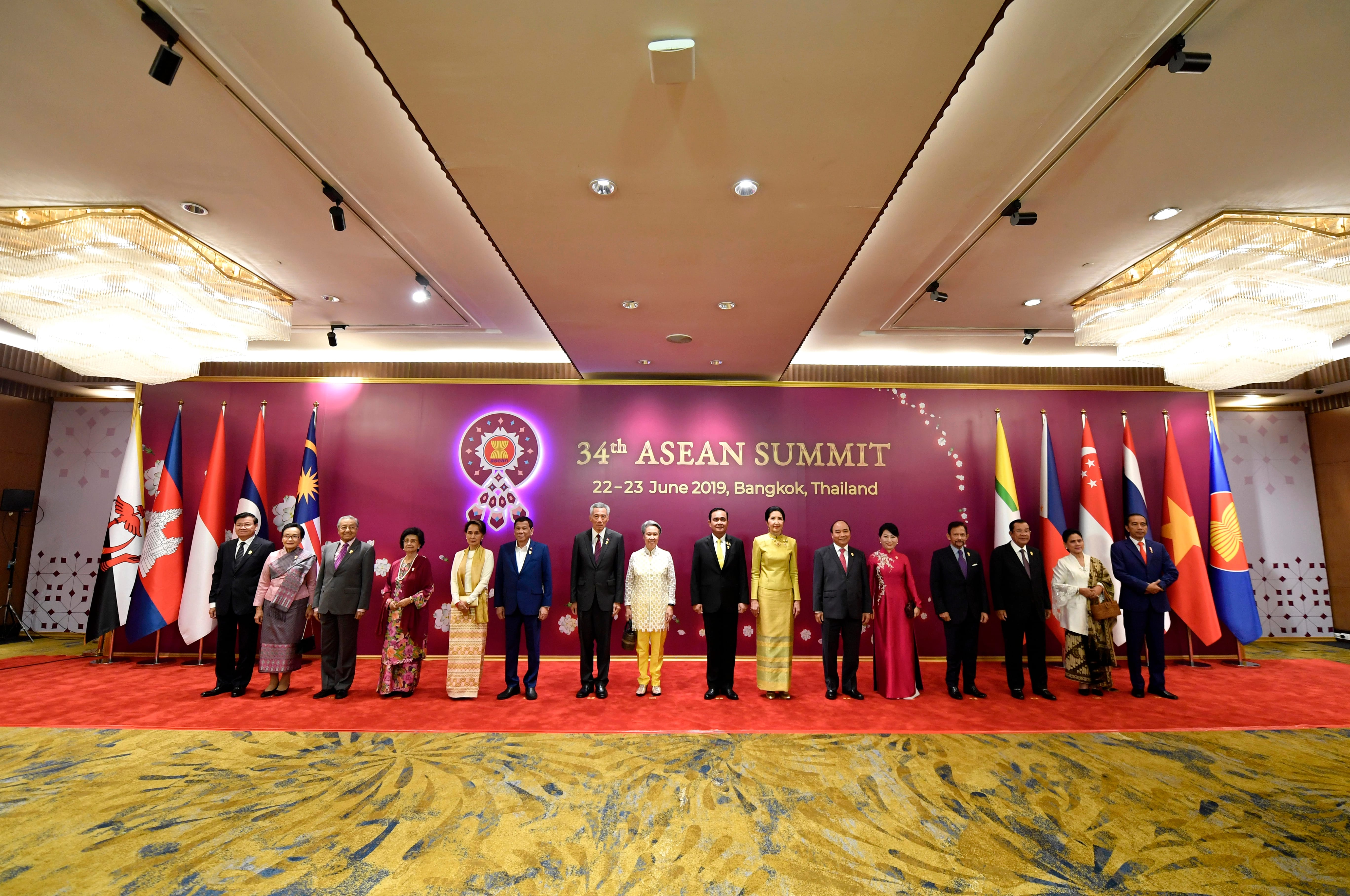 Group posing for "34th ASEAN Summit, Bangkok," with flags, burgundy backdrop and patterned carpet.