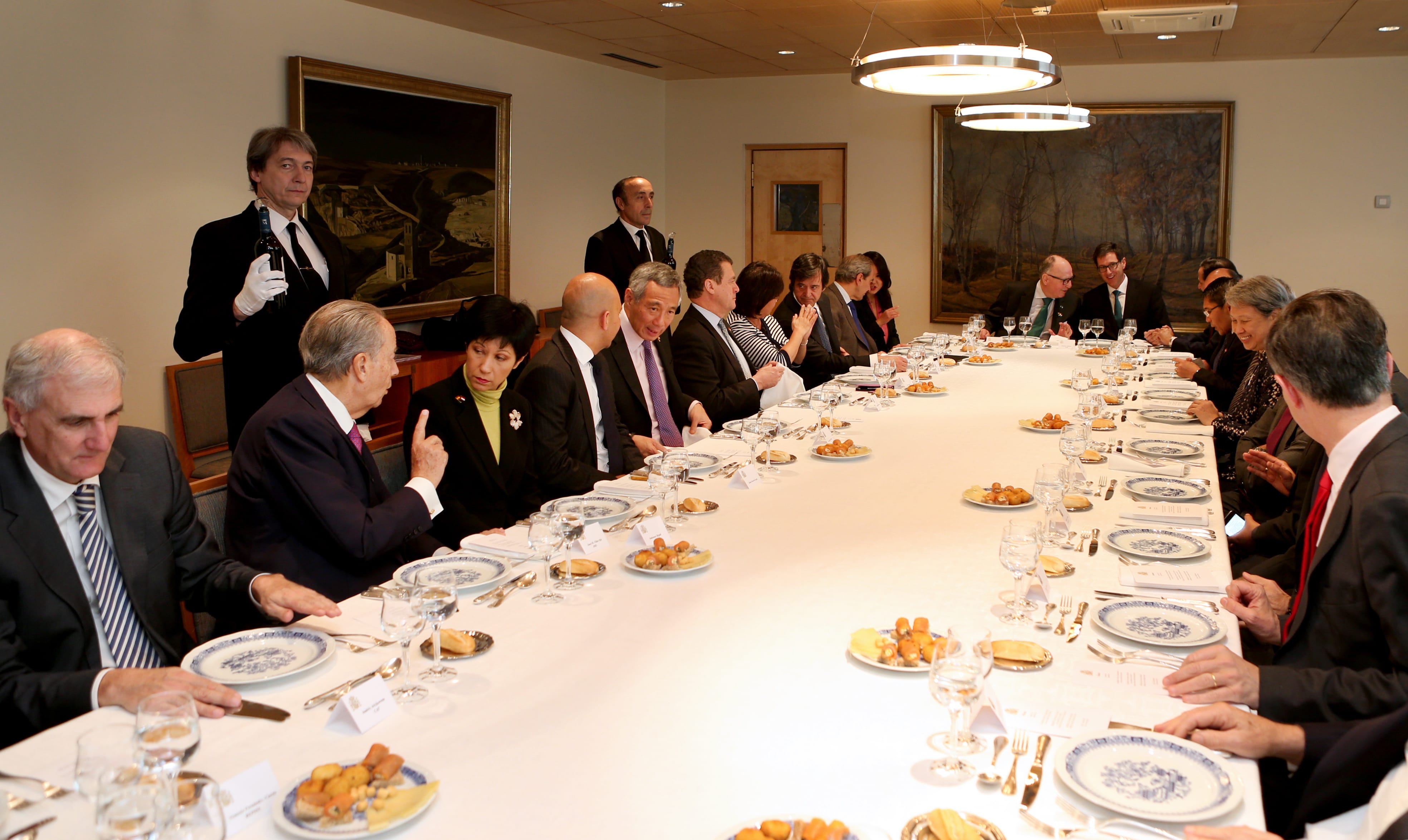 A long table with seated people, set for a meal with servers in black suits standing.