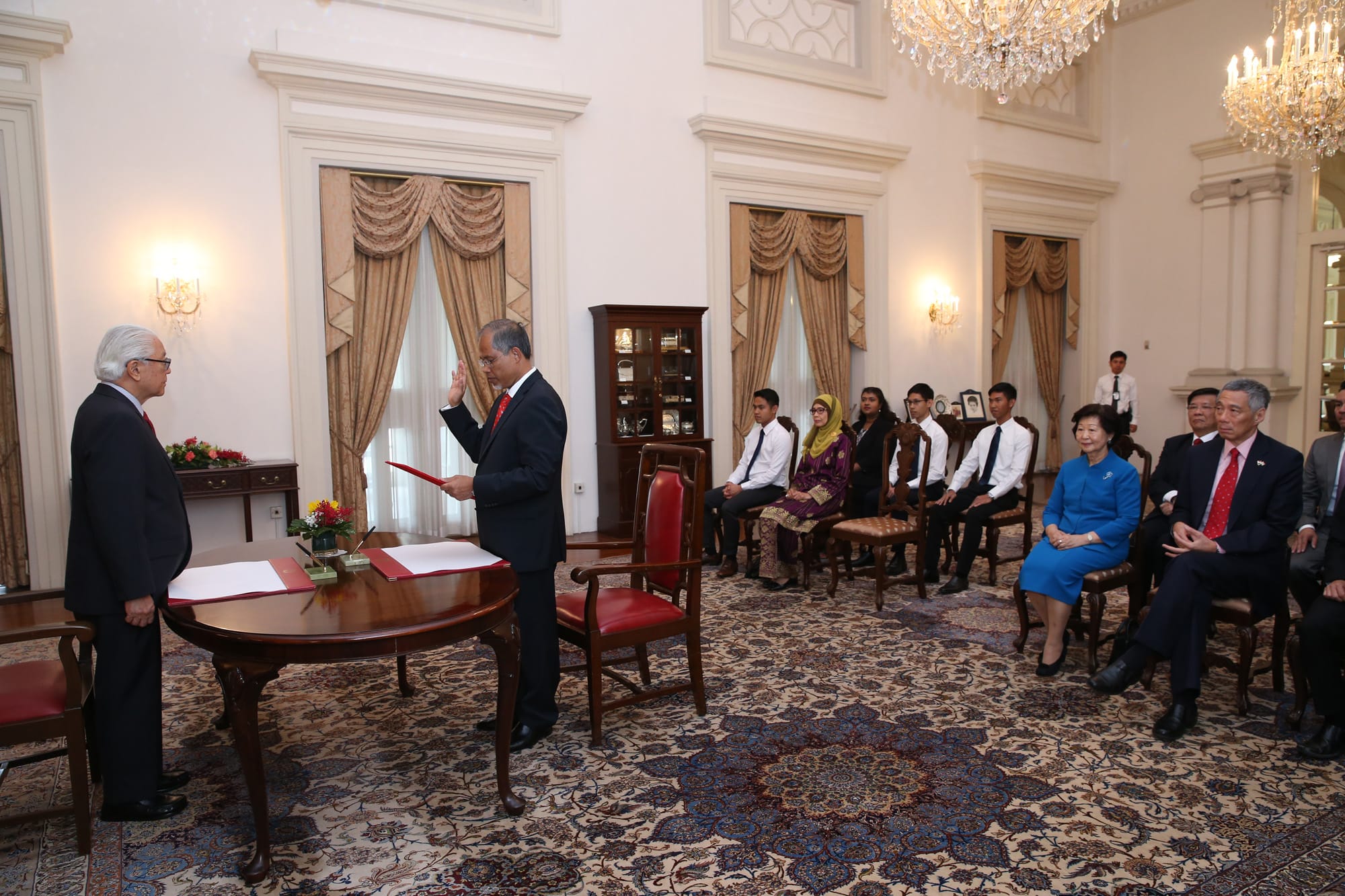 Two men in suits, one with his hand raised, stand at a table before an audience in an ornate room.