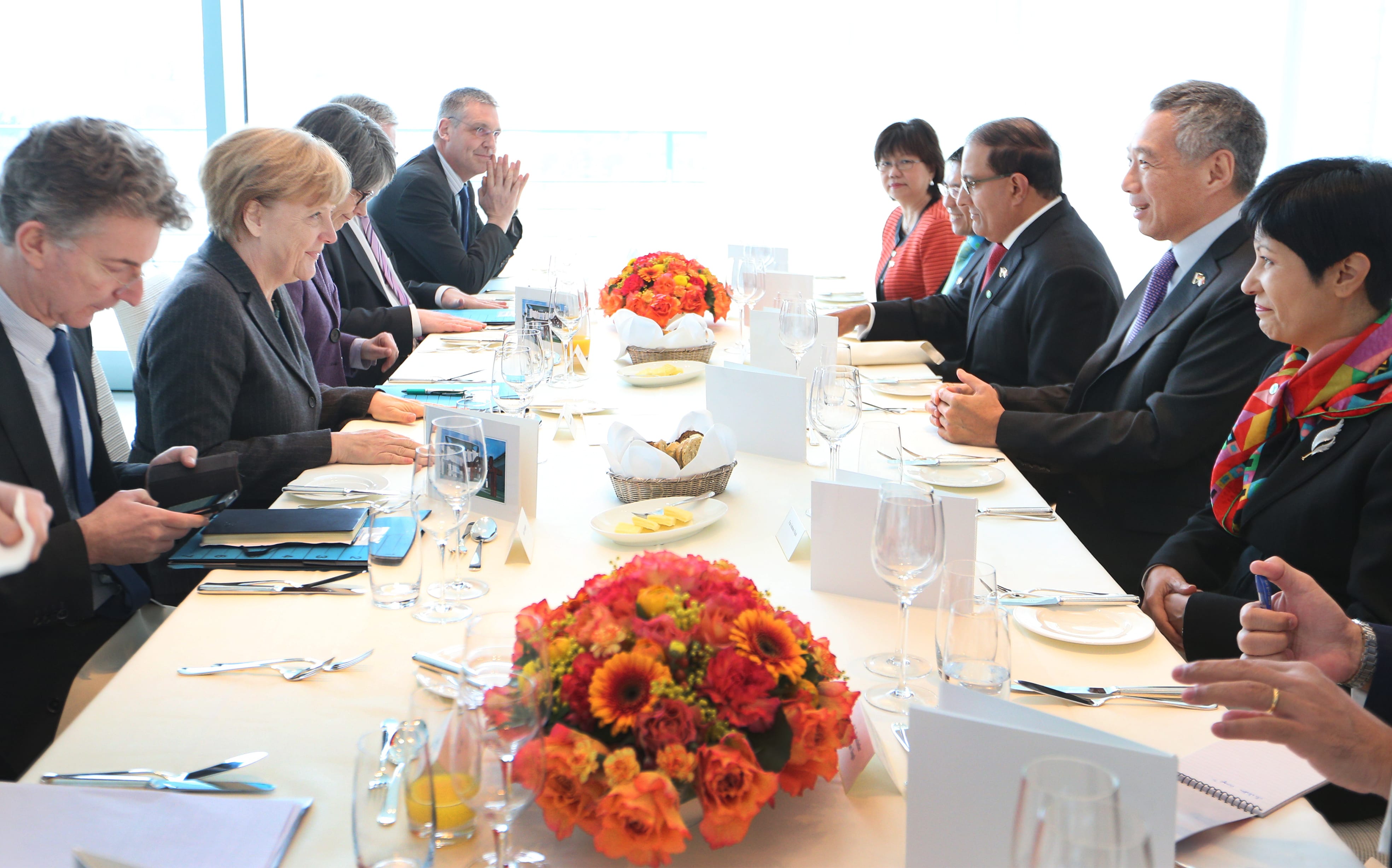 People at a long table, including Angela Merkel and Lee Hsien Loong. Table set with flowers, glasses and tableware.