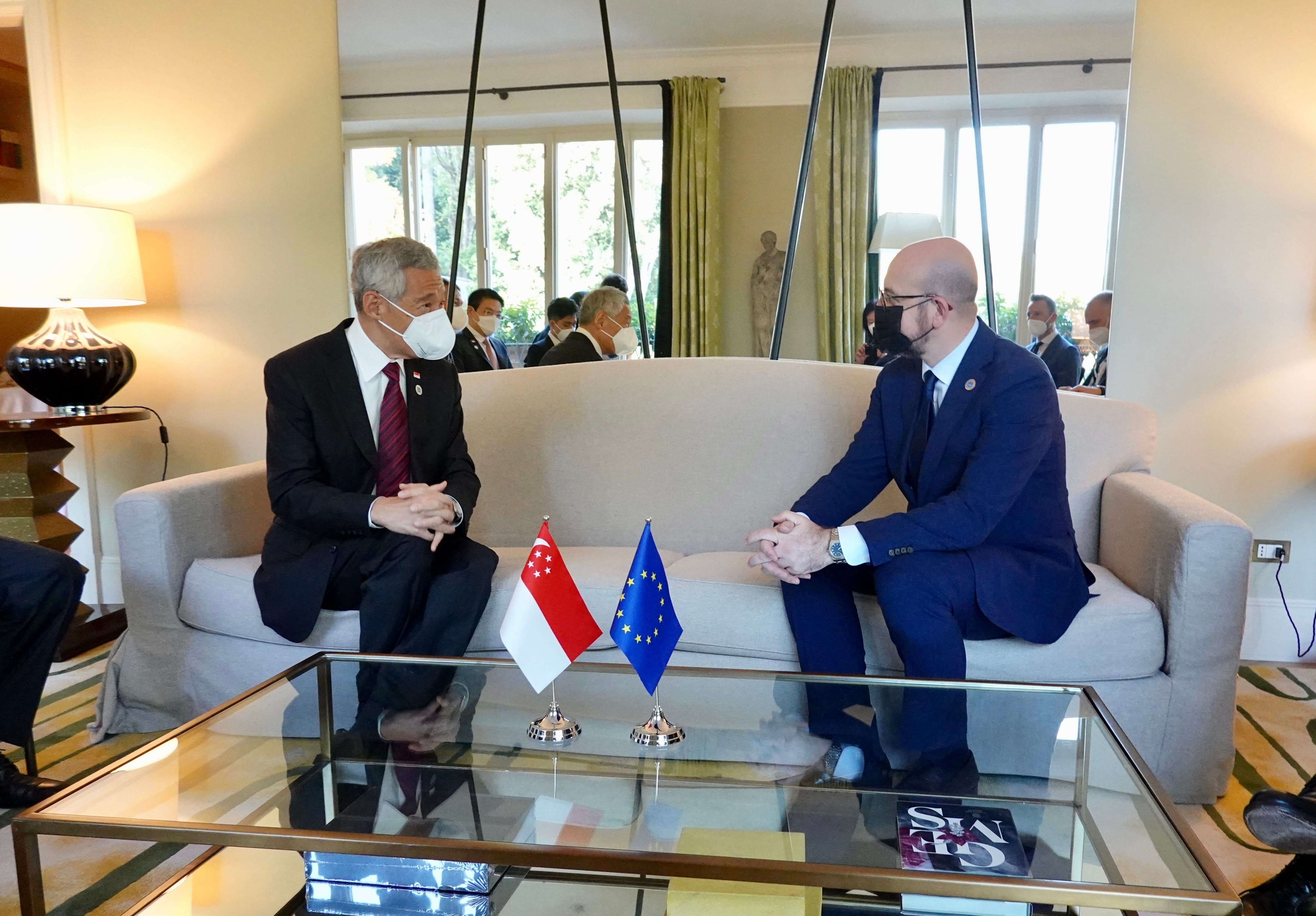 Lee Hsien Loong and Charles Michel sit on a sofa with Singapore and EU flags on a glass table. Both are wearing masks.