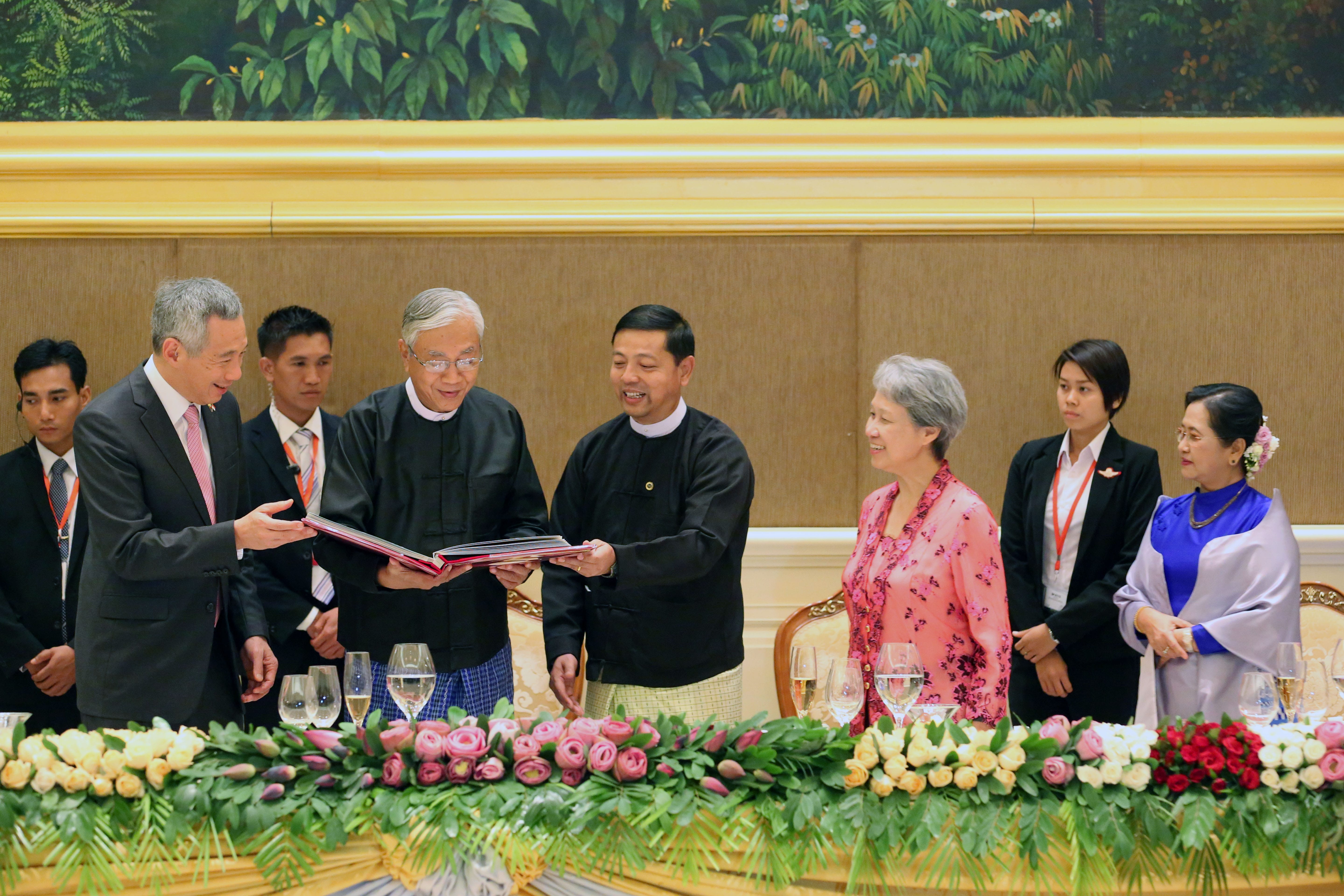 Lee Hsien Loong presenting a book, surrounded by officials and floral arrangements.