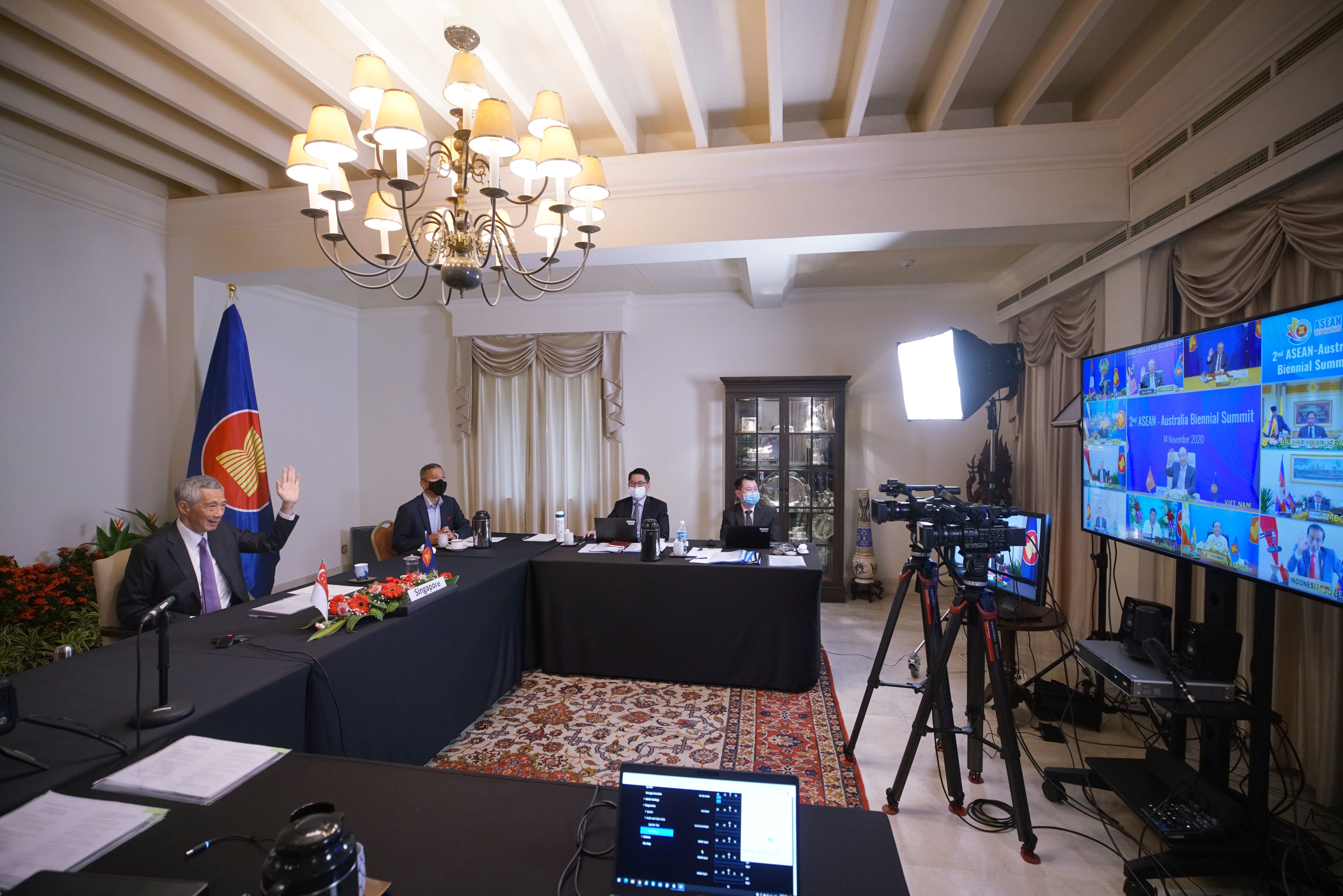 Meeting room showing Lee Hsien Loong with attendees, video cameras, monitors showing video conference.