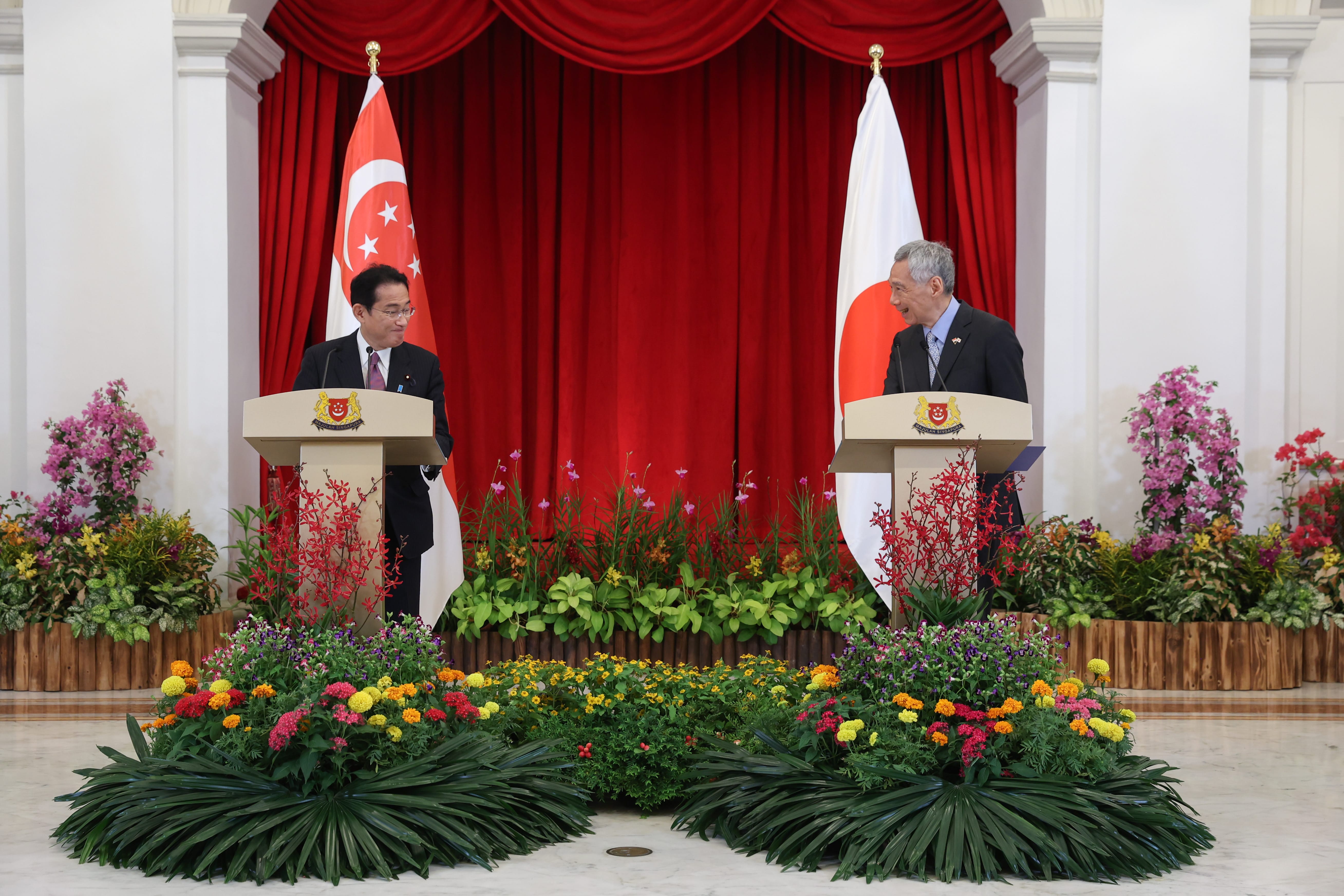 Two men at lecterns with Singapore and Japan flags, colorful flower arrangements decorate the room.