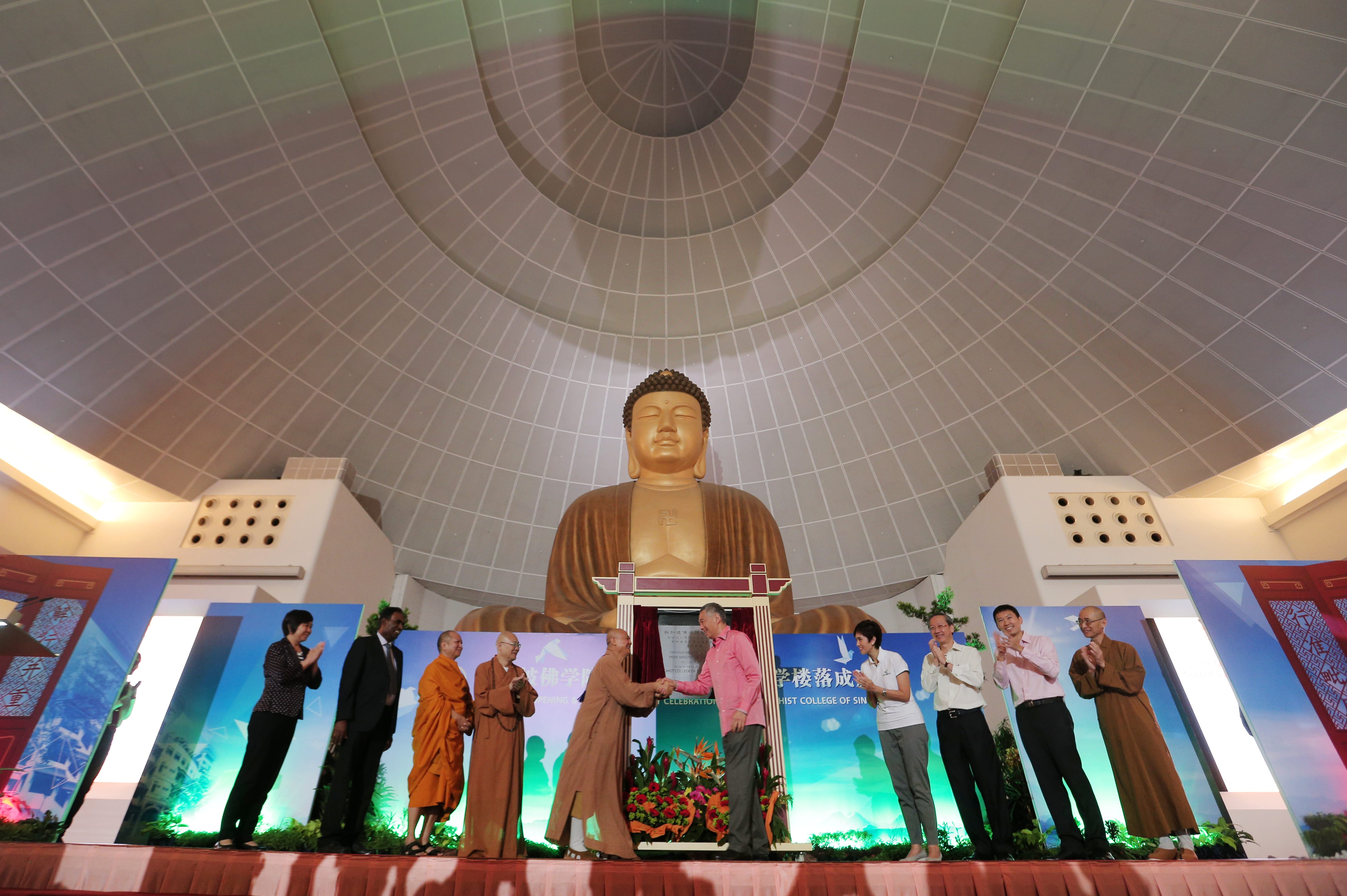 People on stage with large Buddha statue and grid dome. Two people shake hands.