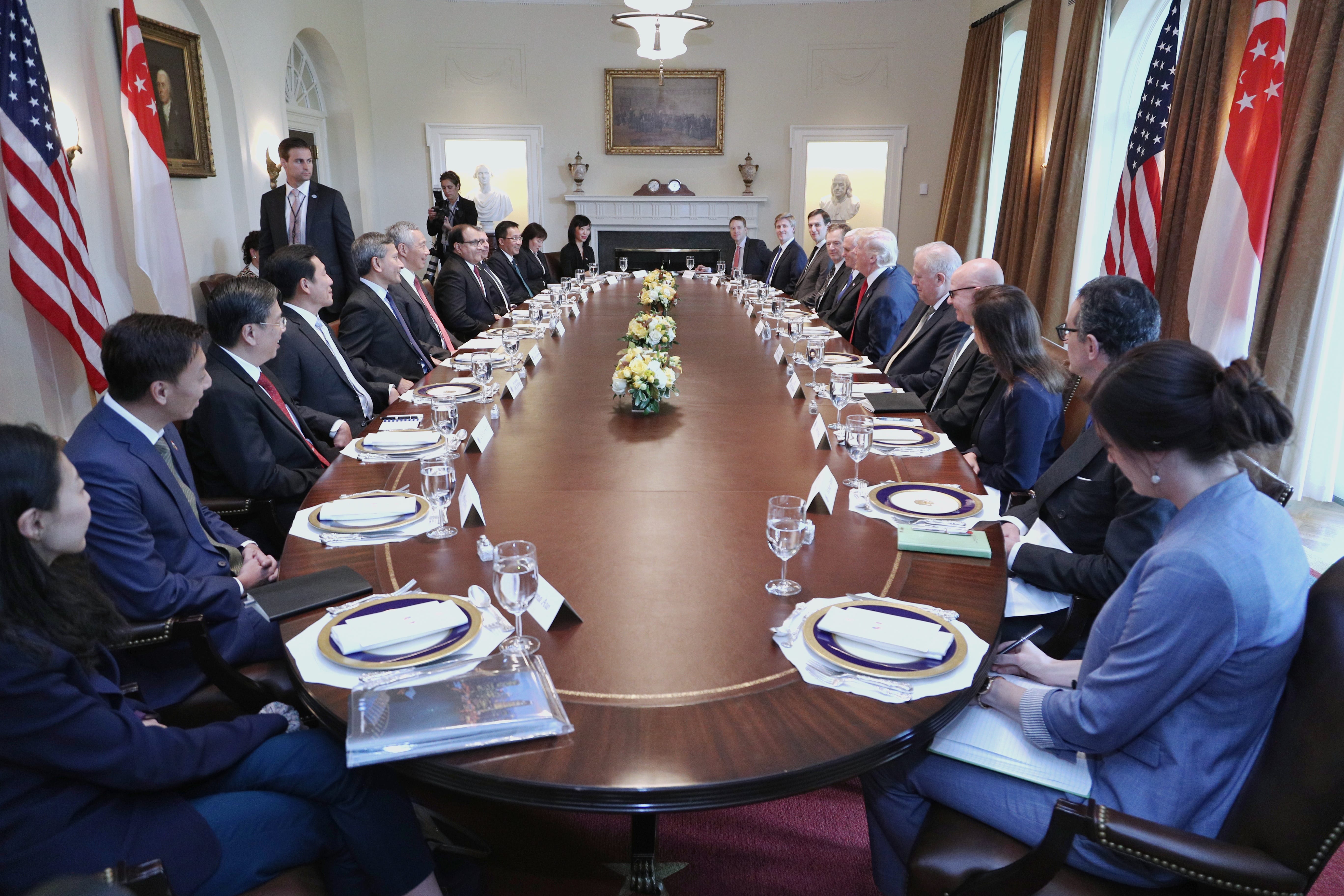 People seated at long table for a meeting with USA and Singapore flags in background.