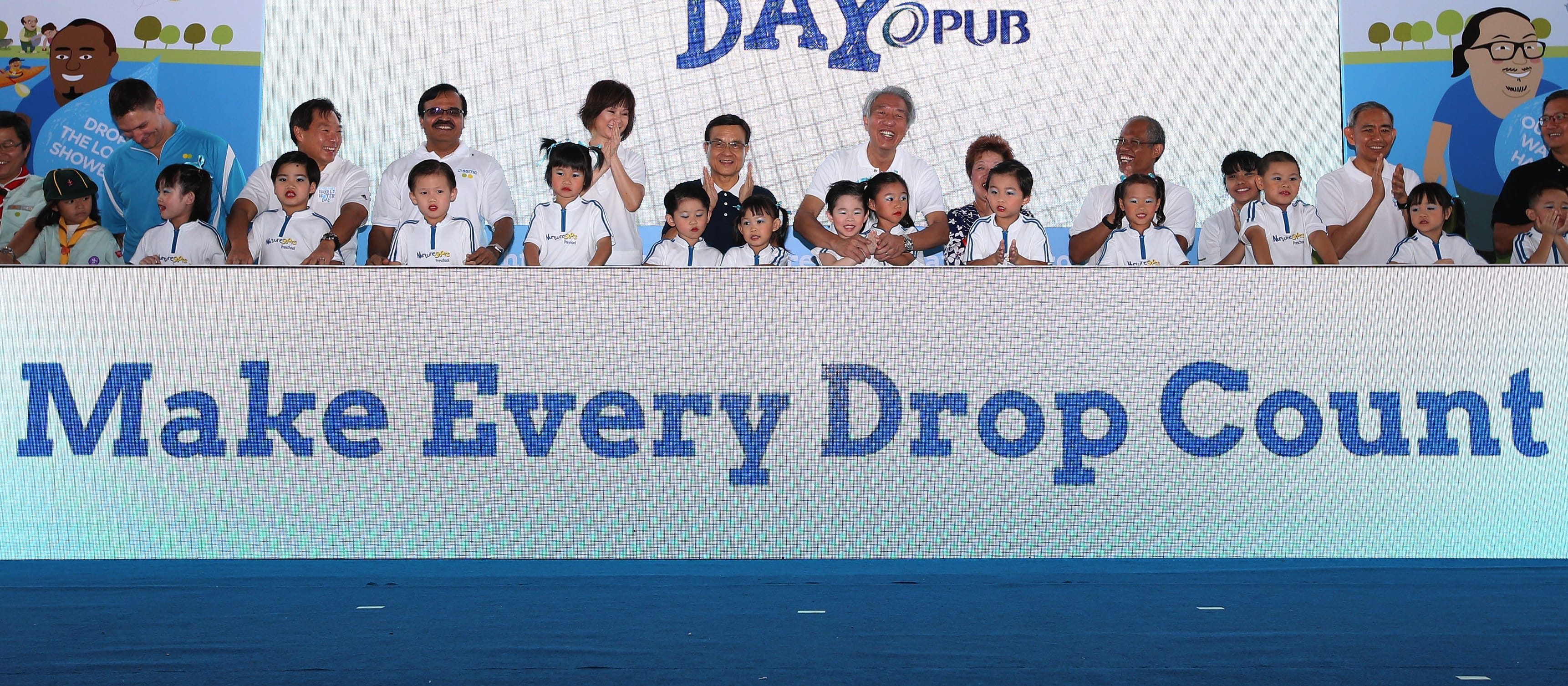 Group of children and adults stand behind a blue screen reading "Make Every Drop Count".