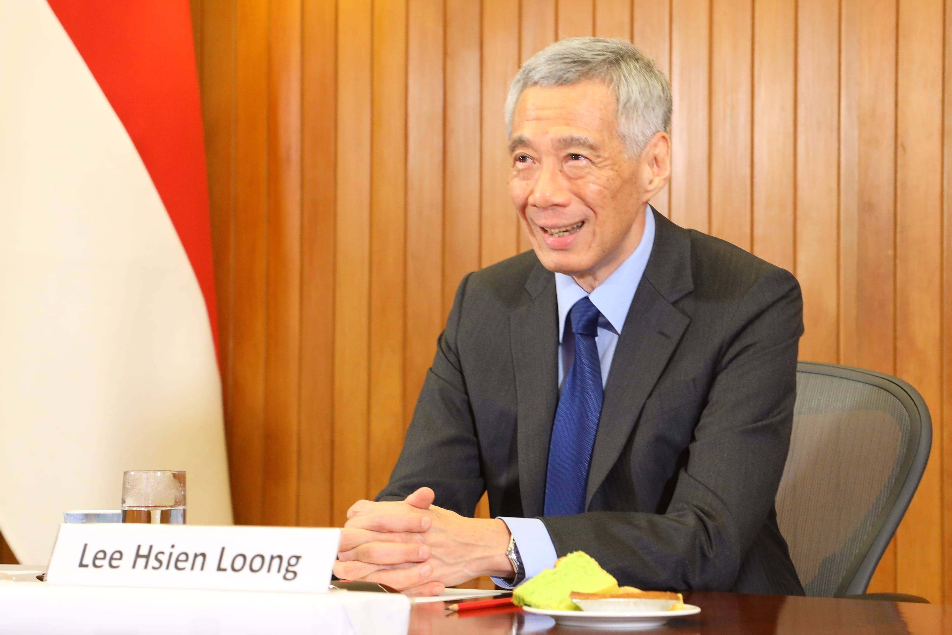 Lee Hsien Loong seated with Singapore flag and wood panel background, wearing suit.