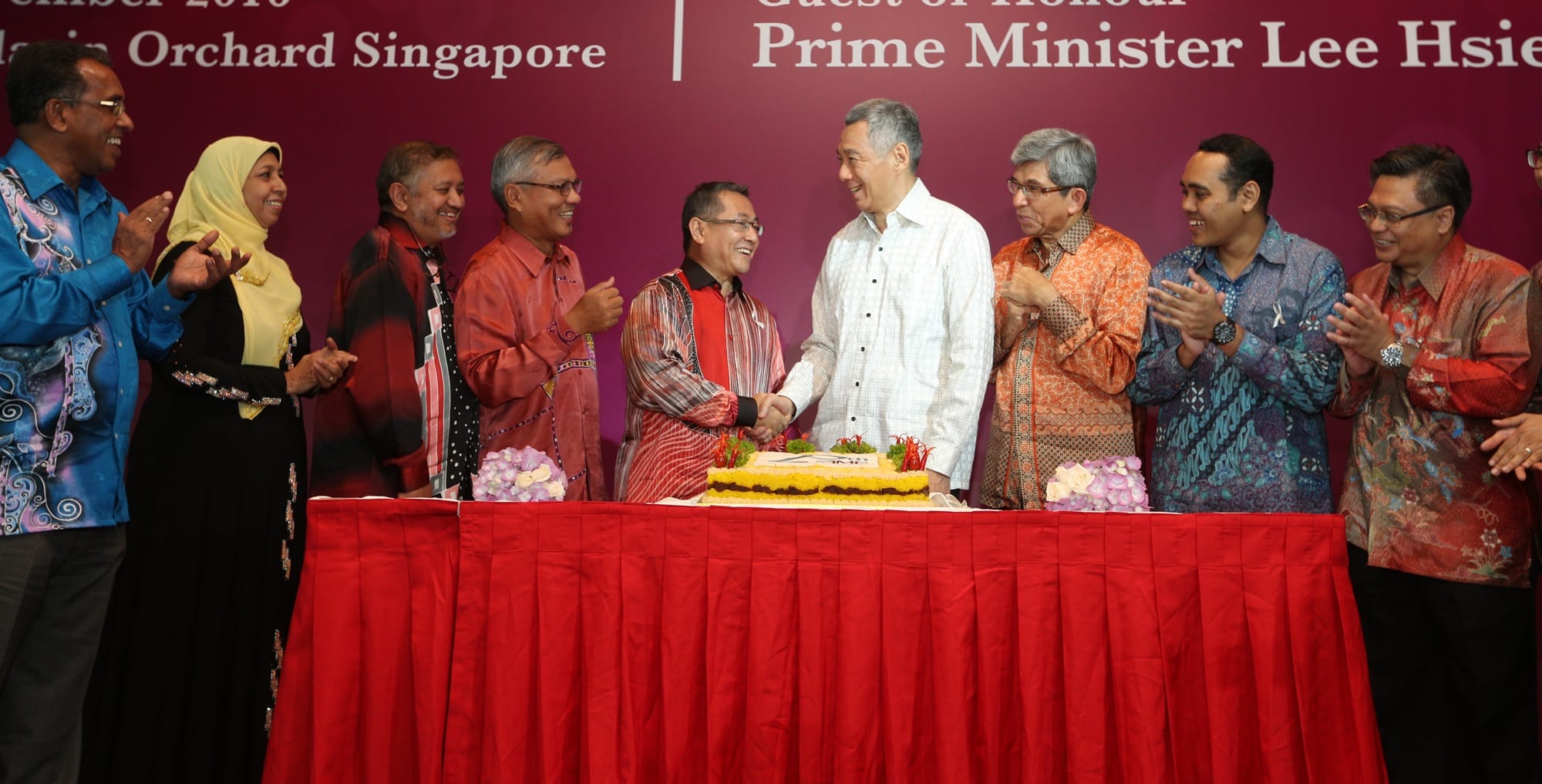 Lee Hsien Loong shakes hands with a man behind a cake, surrounded by people clapping.